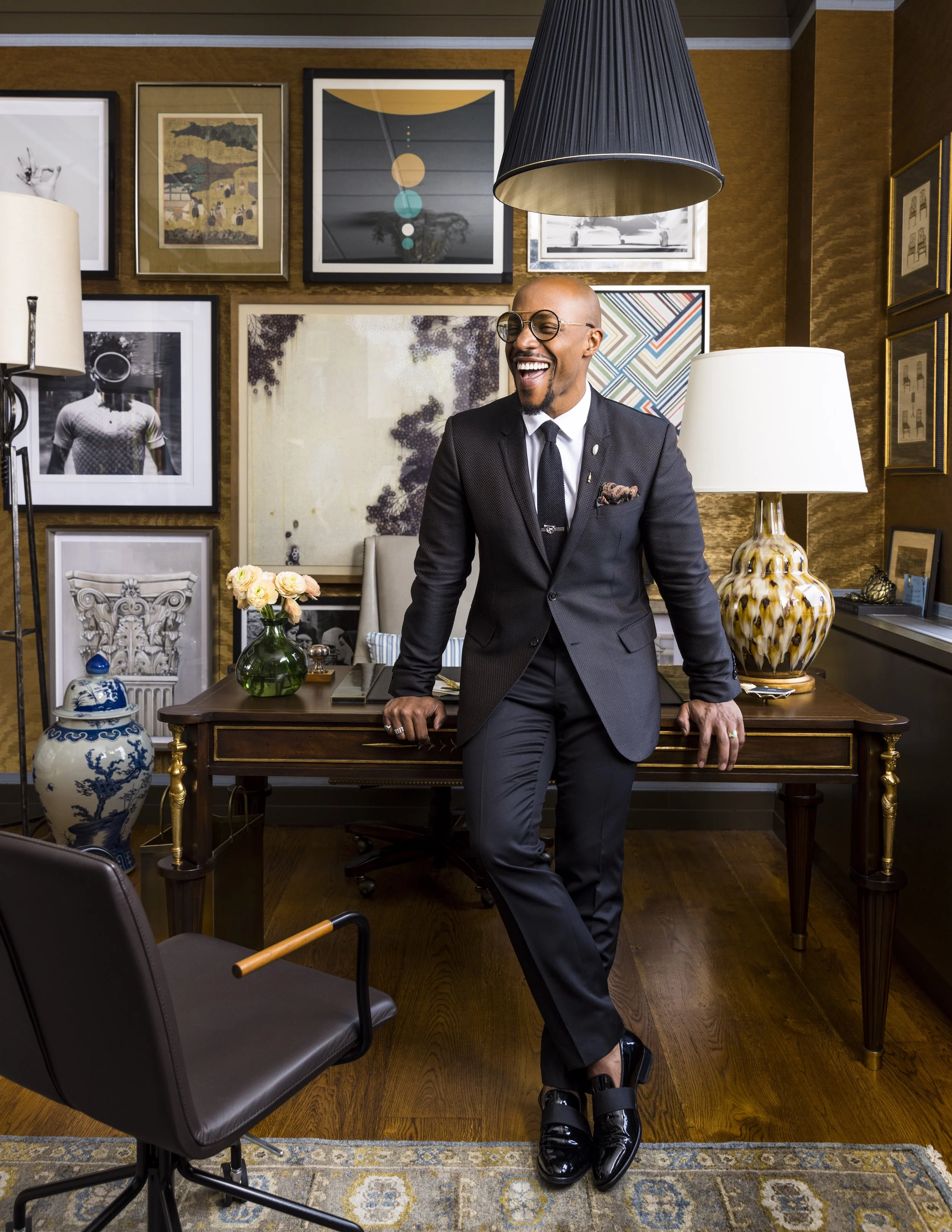 Corey Damen Jenkins in a black suit and glasses, smiling and leaning on a desk in an office decorated with framed artwork and various lamps.