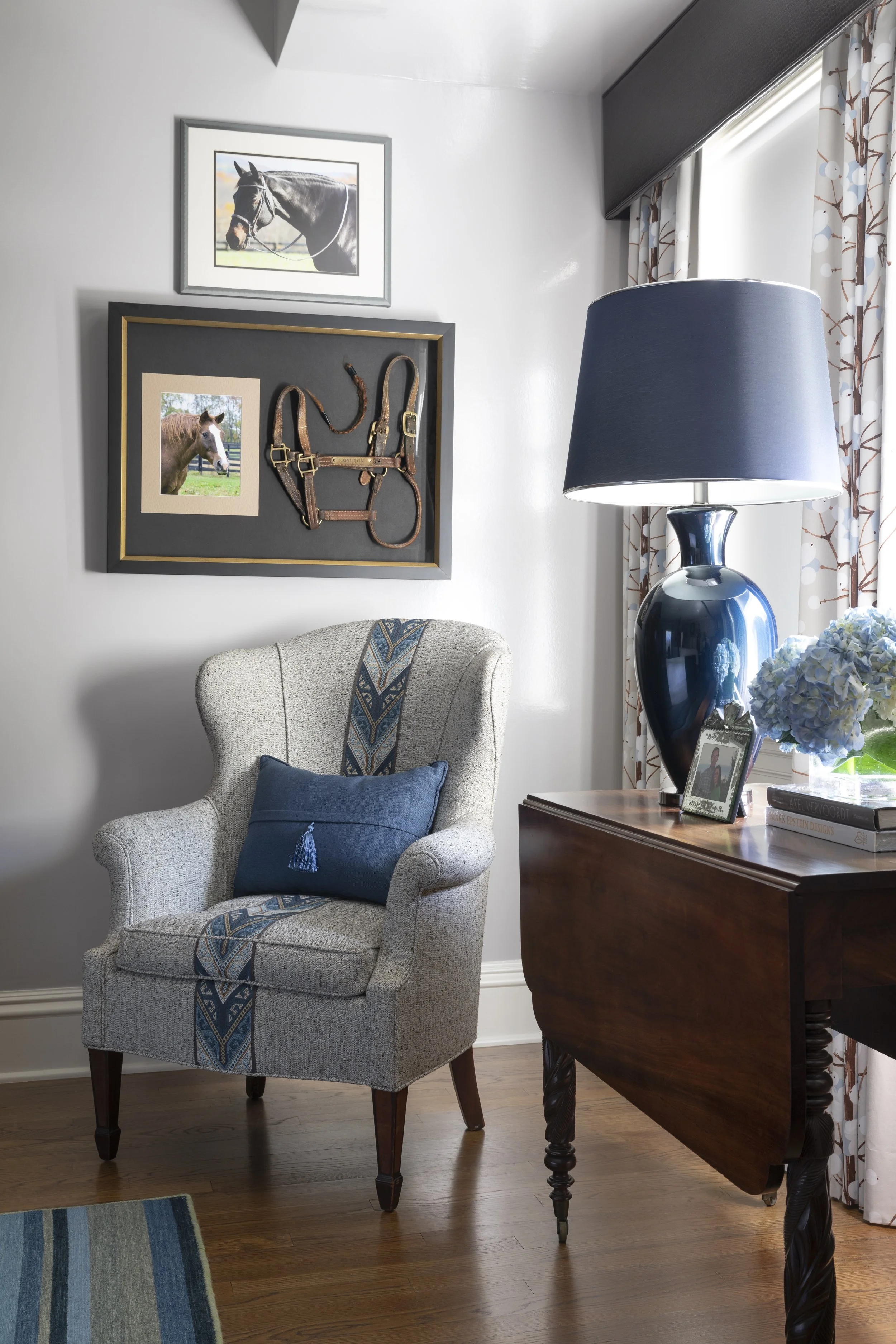 A cozy living room corner with a cream upholstered armchair, a dark wood side table, a large blue ceramic table lamp, and a window with white and brown patterned curtains. Two framed pictures of horses hang on the white wall above the armchair, along