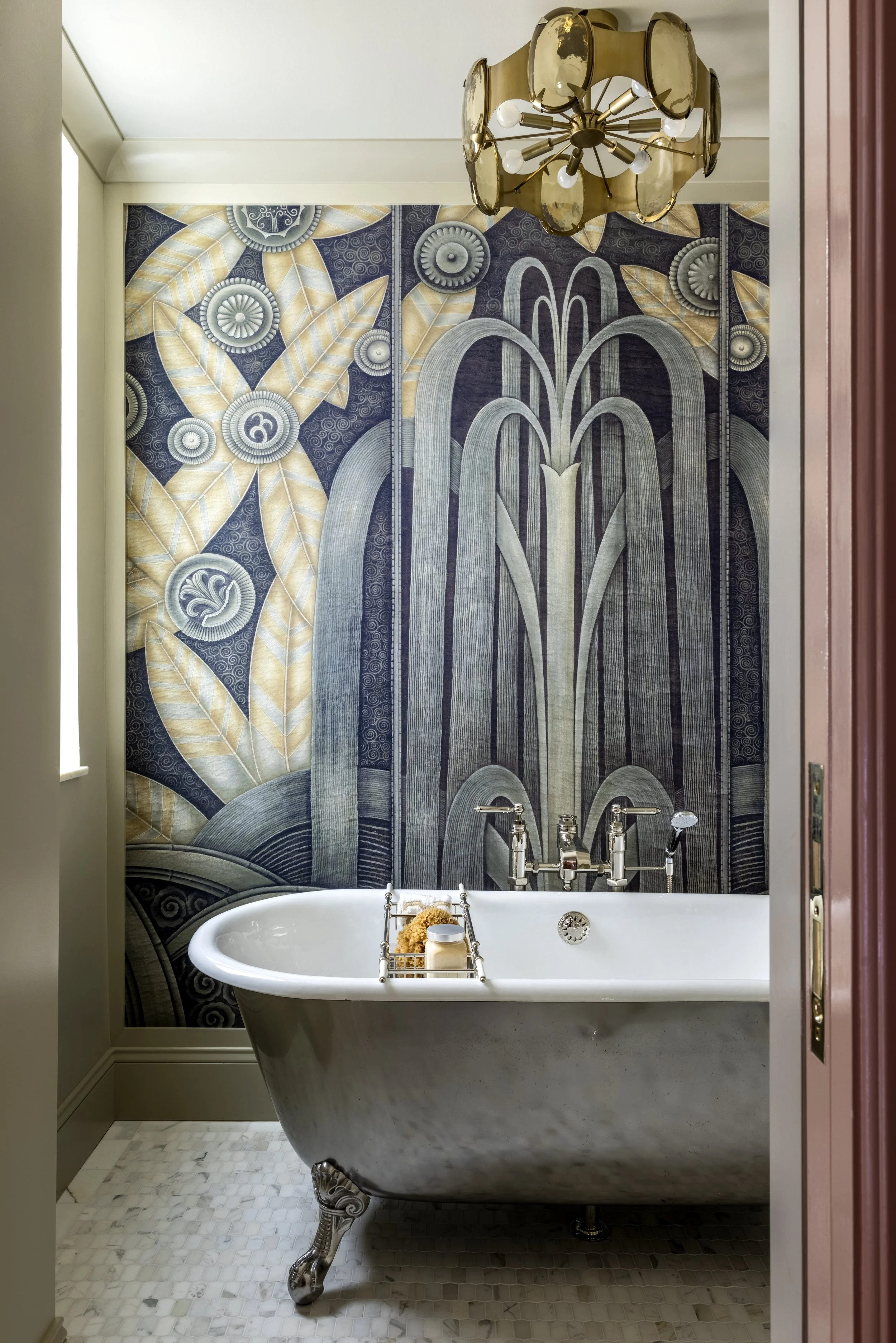 A vintage clawfoot bathtub in a bathroom with a decorative mural of stylized fountains and leaves on the wall behind it. The bathtub has ornate metal feet and a tray holding toiletries. A modern chandelier hangs from the ceiling.