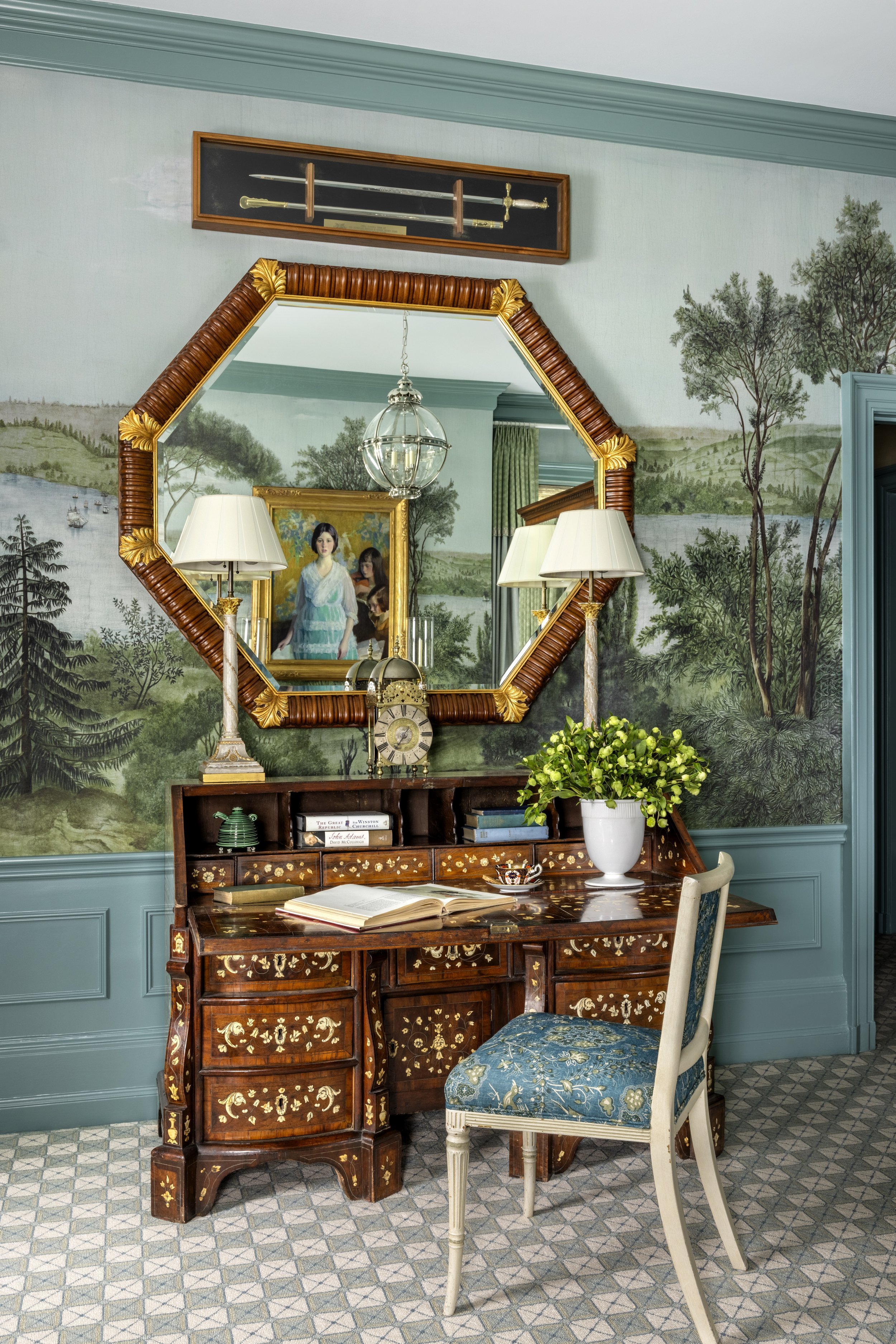 A room with a large leafy mural wallpaper, a wooden desk with intricate inlay design, an open book, and a white chair with blue patterned upholstery. There is a large mirror with a decorative frame, two lamps, a potted plant, and a framed sword displ