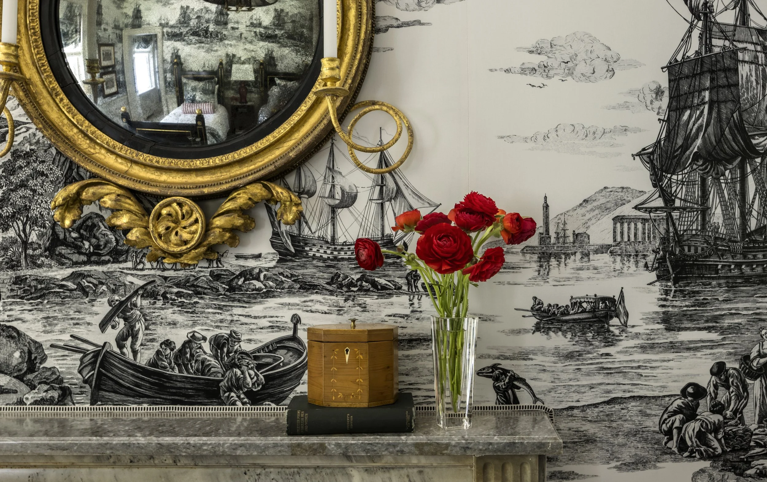 A decorative wall with black and white nautical scene mural, a gold ornate round mirror, a bouquet of red flowers in a clear glass vase, a wooden box, and books on a marble surface.