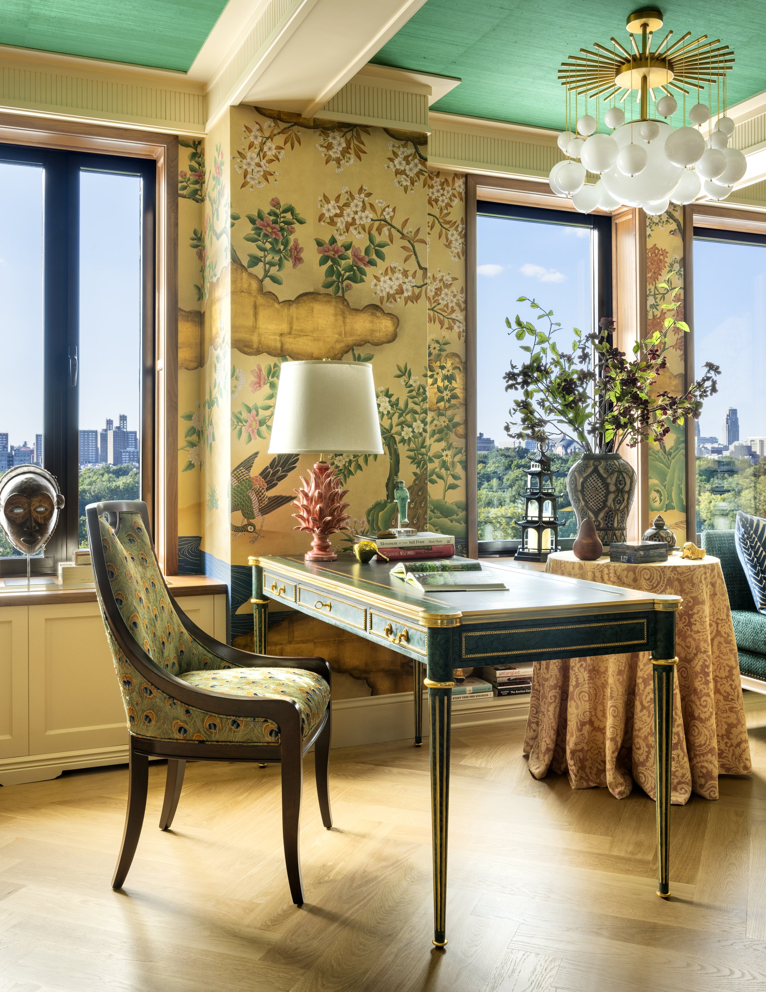 A sunlit room with large windows showing a cityscape of tall buildings. Inside, there is a green and gold desk with books and a decorative pink lamp. A patterned upholstered chair is nearby, and a round table with a floral tablecloth holds a large va