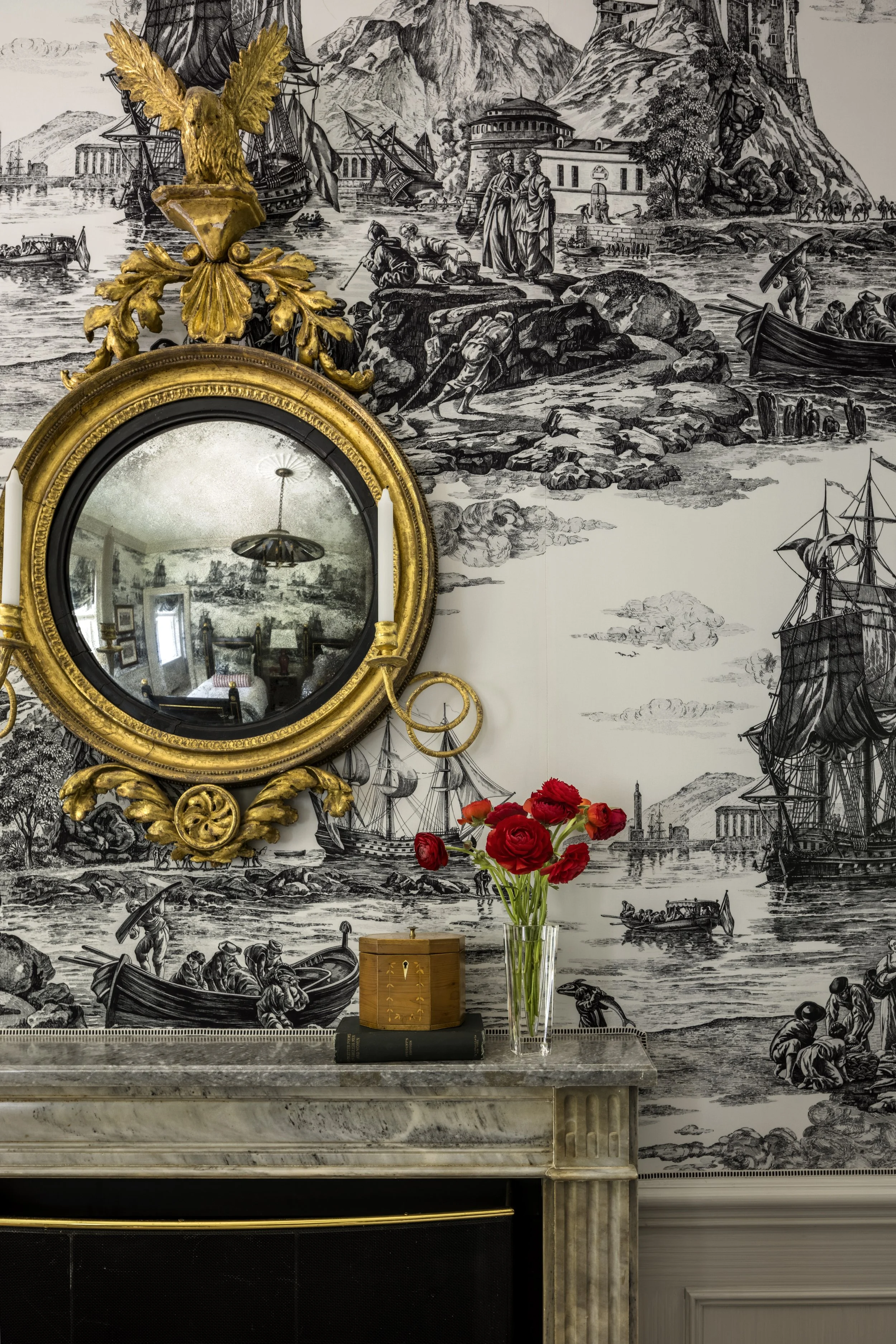 A decorative room with a marble fireplace, a round ornate gold-framed mirror, a vase of red roses, and a black and white mural depicting ships, mountains, and people in historical attire.