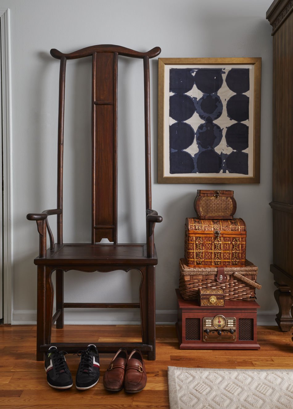A hallway corner with a dark wooden antique hall chair, a pair of black and white sneakers, brown loafers, a wicker basket stack, a vintage radio, a framed blue and white abstract art, and a light-colored rug on a hardwood floor.