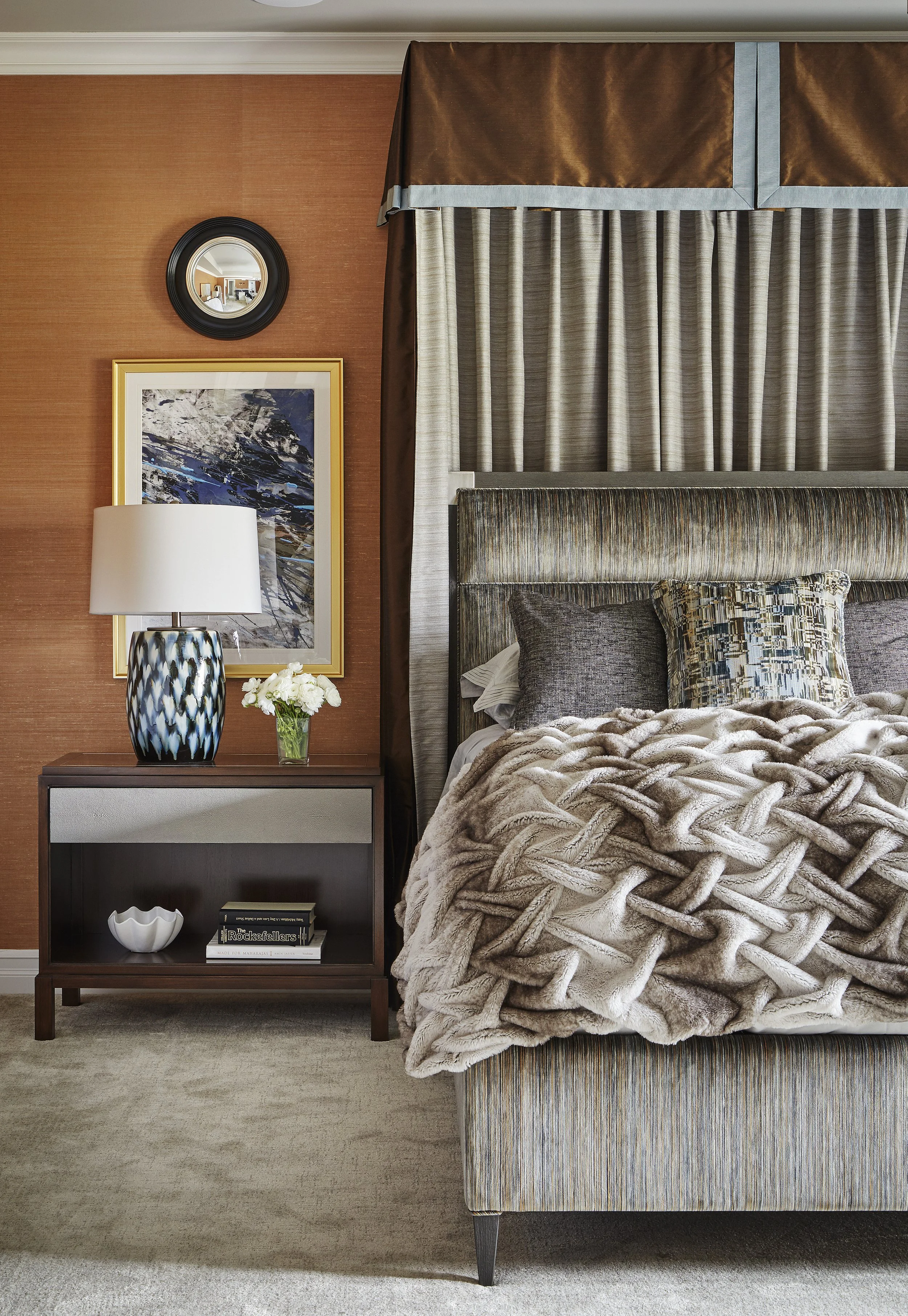 A bedroom corner with a bed, pillows, headboard, curtains, and a side table holding a lamp, framed artwork, and a small vase with flowers.