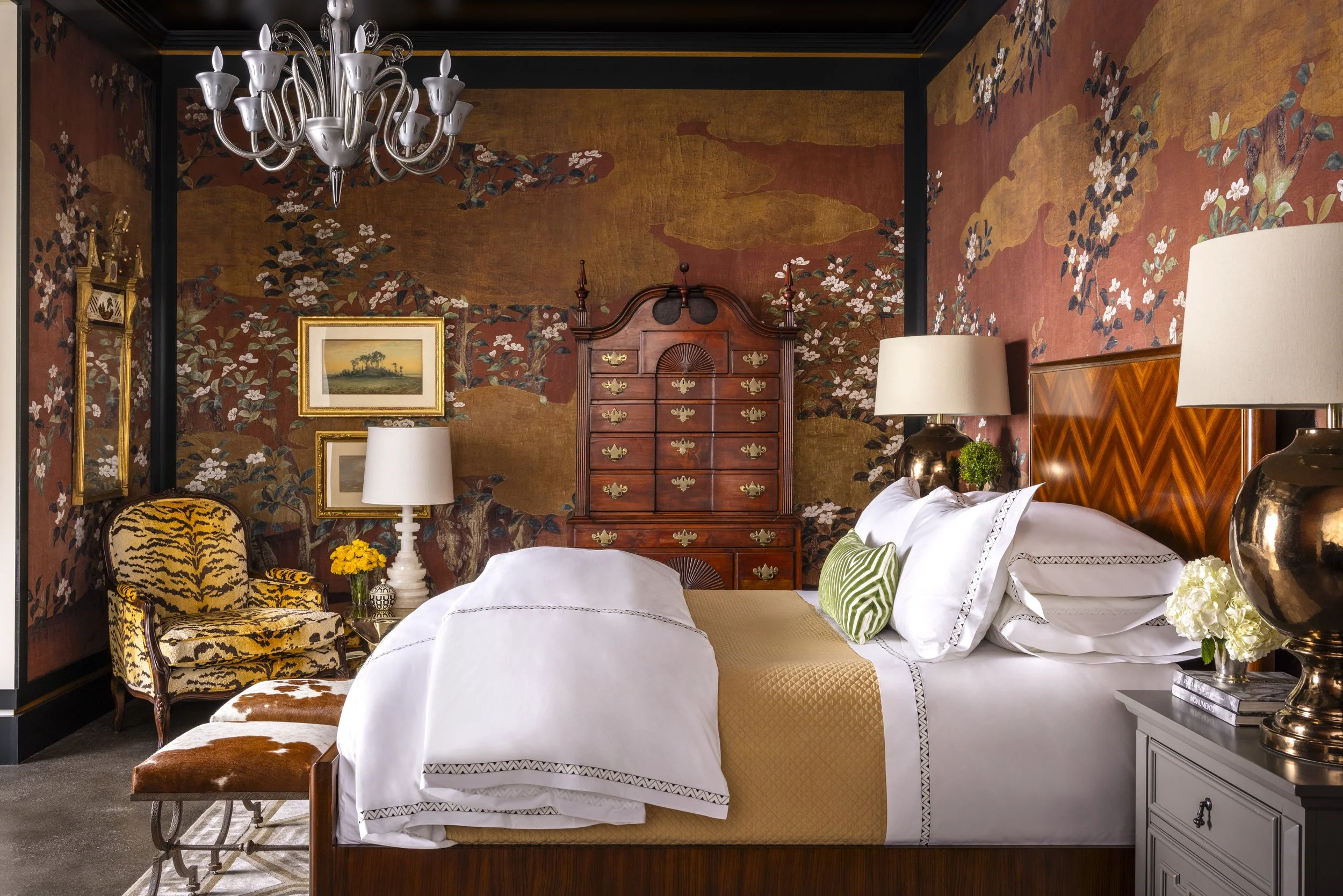 A bedroom featuring a vintage-style patterned wall, a wooden dresser with brass hardware, a bed with white and green bedding, lamps, framed artwork, a chandelier, a tiger print chair, and various decorative items.