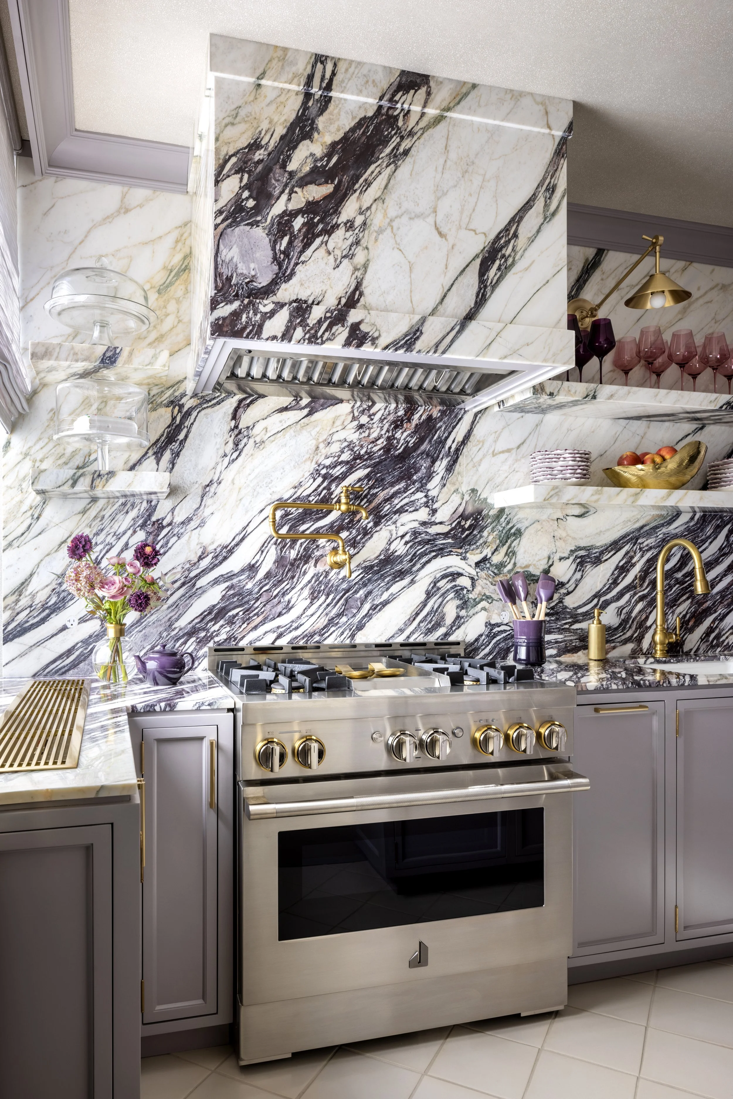 Modern kitchen with marble countertops and backsplash, gray cabinets with gold handles, stainless steel oven, gold faucet, pink and purple wine glasses, flowers, and decorative gold bowl with apples.