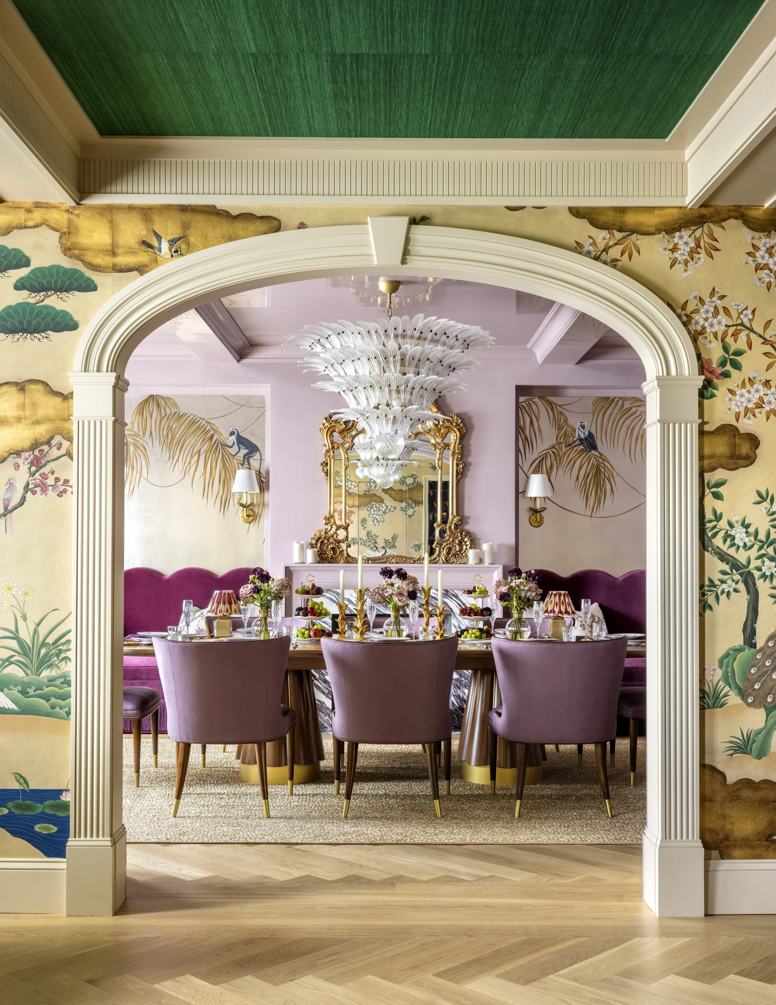 Elegant dining room with a pink velvet banquette, a round wooden table, purple velvet chairs, and a large ornate chandelier. The room features floral and nature-themed wallpaper, decorative wall sconces, and a mirror with a gilded frame.
