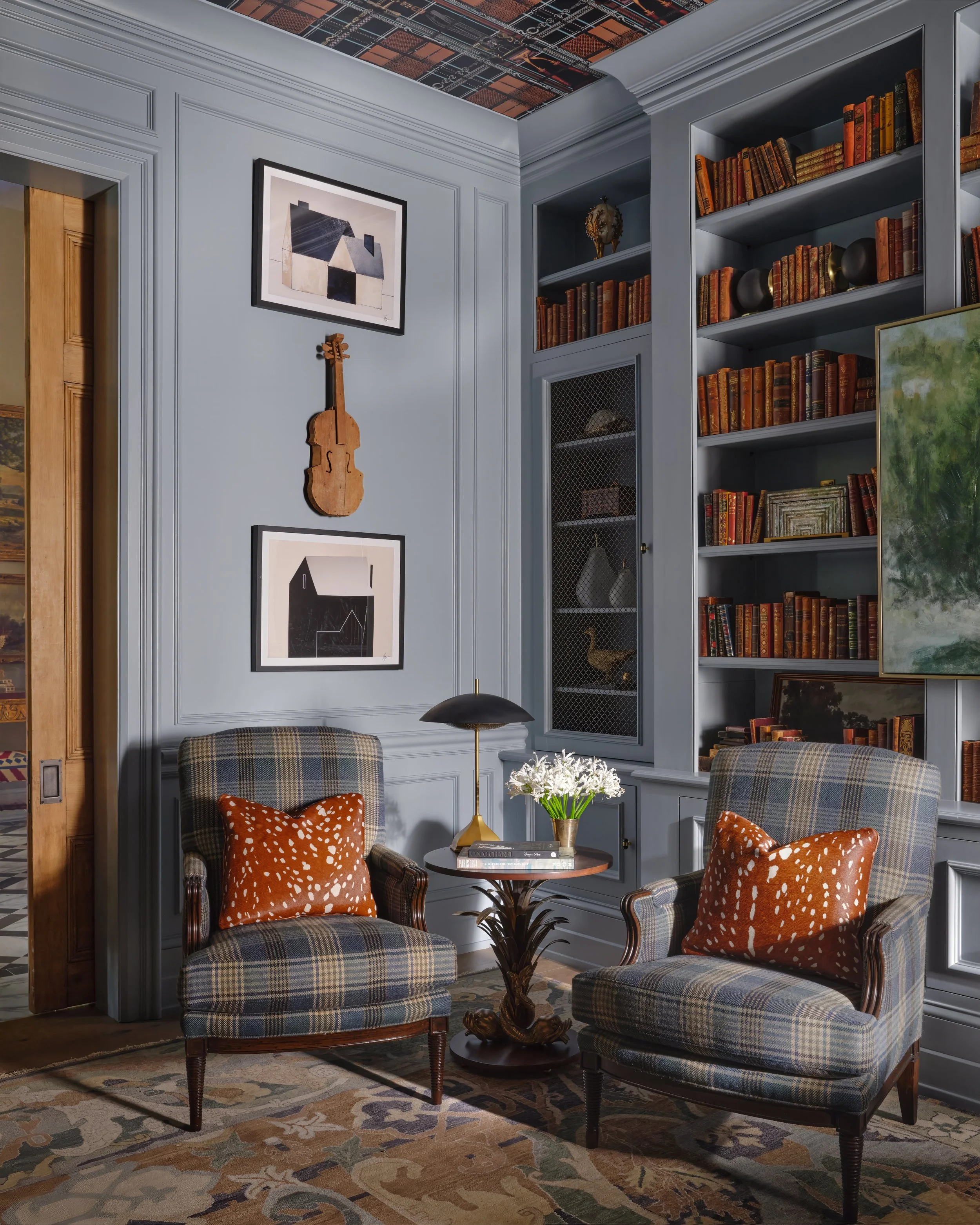 A cozy sitting area with two plaid armchairs with orange patterned pillows, a round wooden table with books and a decorative pineapple, a black and gold table lamp, a wall with blue paneling, framed artwork, a small wooden violin hanging on the wall,