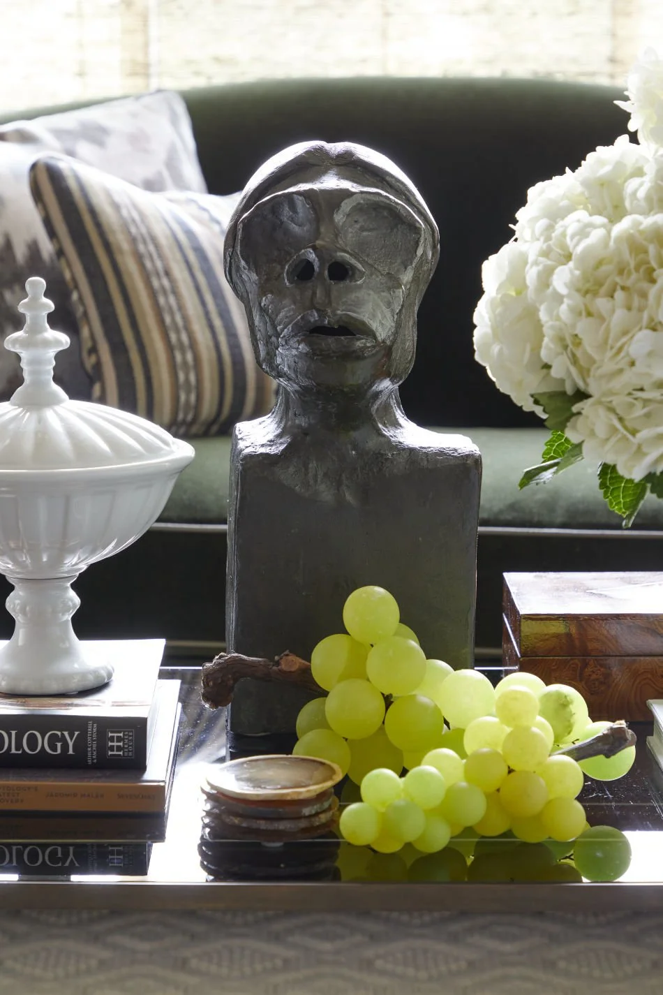 A decorative table setting with a black abstract sculpture of a human face, a bunch of green grapes, a white ceramic container, a stack of shells, a volume of books, flowers, and a striped pillow in the background.