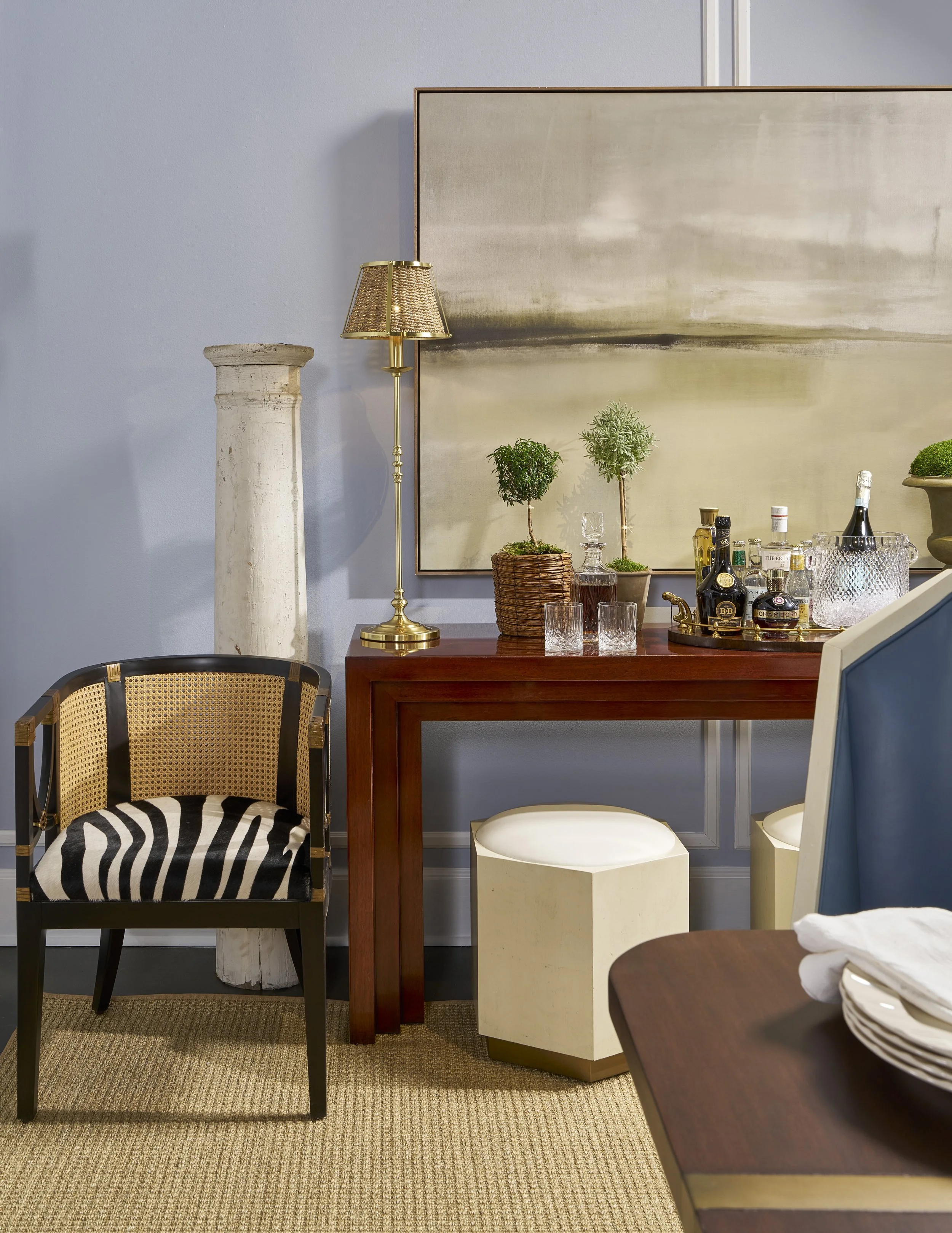 A corner in a room with a bar table, various alcohol bottles, a gold lamp, a pair of small potted plants, and a large abstract painting on the wall. There are chairs and a smaller table with stacked plates.