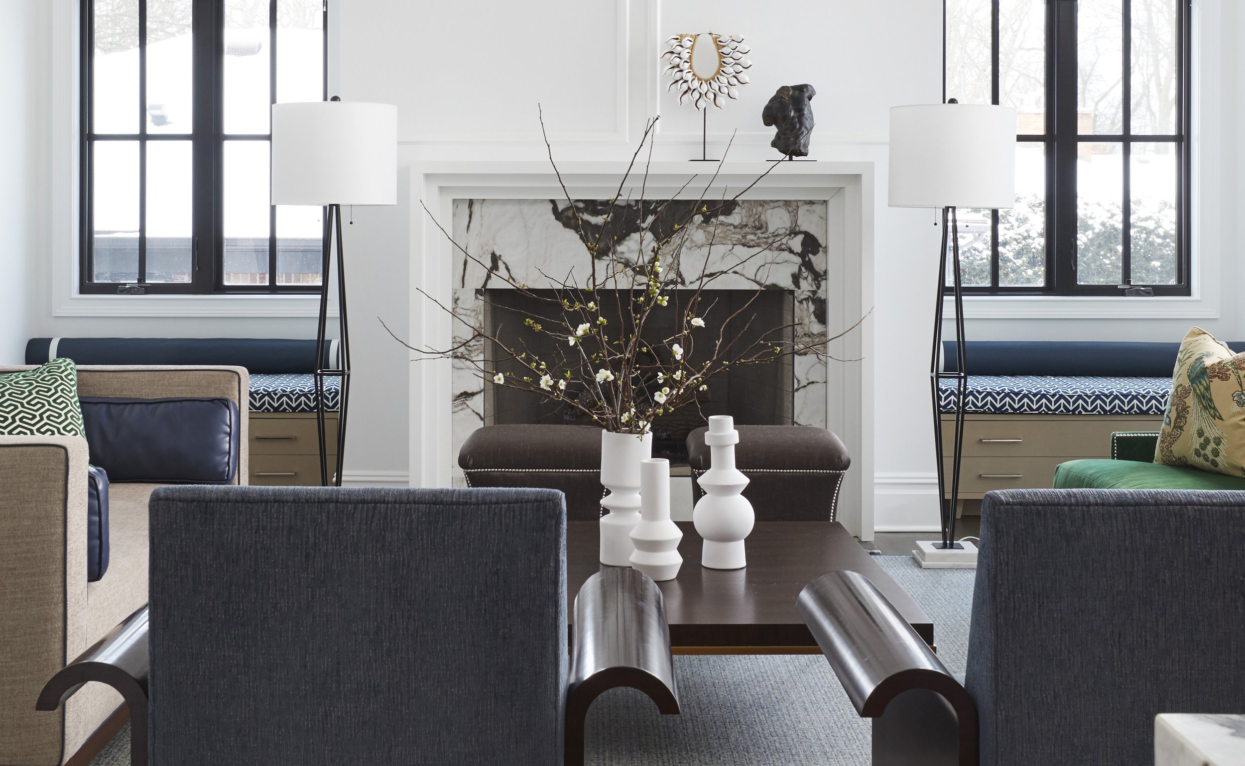 Living room with two large windows, fireplace with marble surround, decorative objects on mantel, tall white lamps on either side of fireplace, a branch arrangement with white flowers in a white vase on a wooden table, and various chairs and cushions