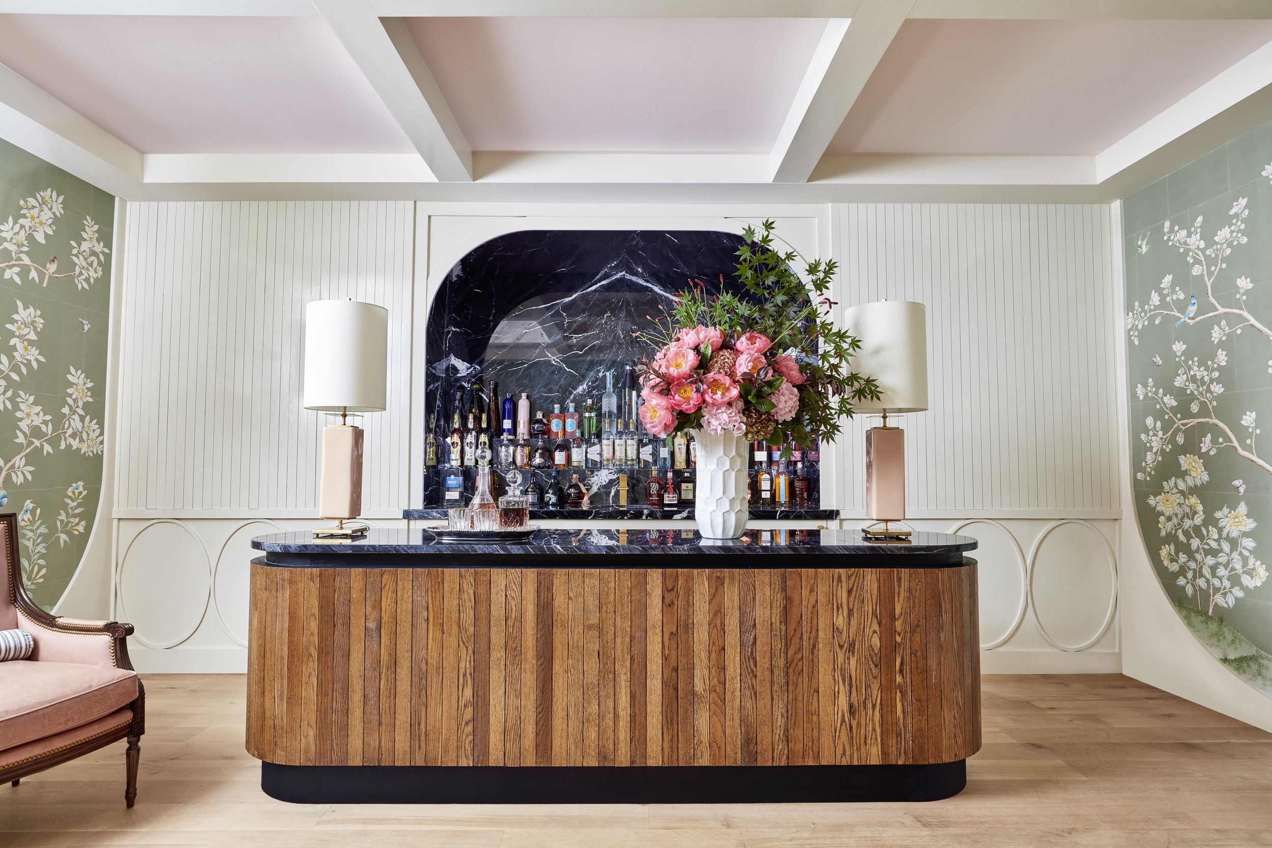 A stylish bar area with a black marble countertop, a large white vase with pink flowers, two white lamps, and various bottles of alcohol. The back wall features a black marble arch and decorative green floral wallpaper on the sides, with wood floorin