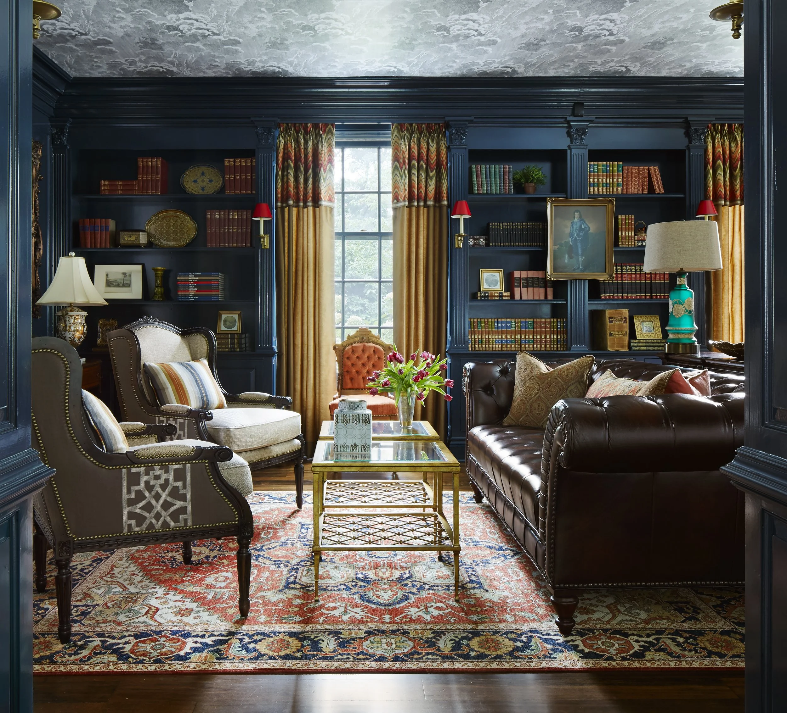 Living room with navy blue built-in bookshelves, gold and red window curtains, a large window, traditional furniture including an armchair, leather sofa, floral ottoman, and rug, decorative lamps, framed photos and artwork, and a bouquet of pink flow