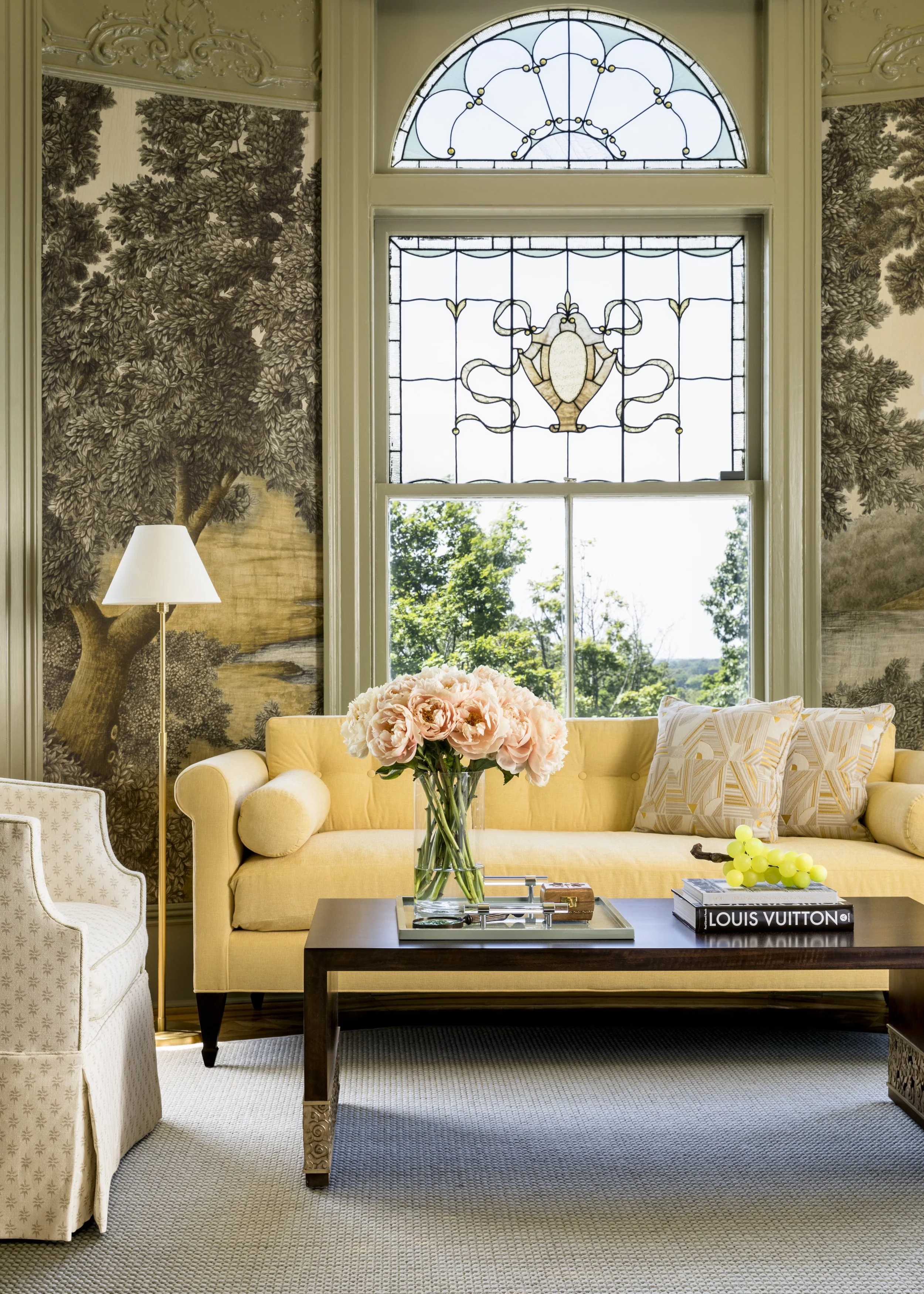 Living room with a yellow sofa, floral pillows, a glass vase with pink peonies on a dark wooden coffee table, a Louis Vuitton book, a bunch of grapes, and a gold lamp. The back wall features detailed tree wallpaper, and a large window with stained gl