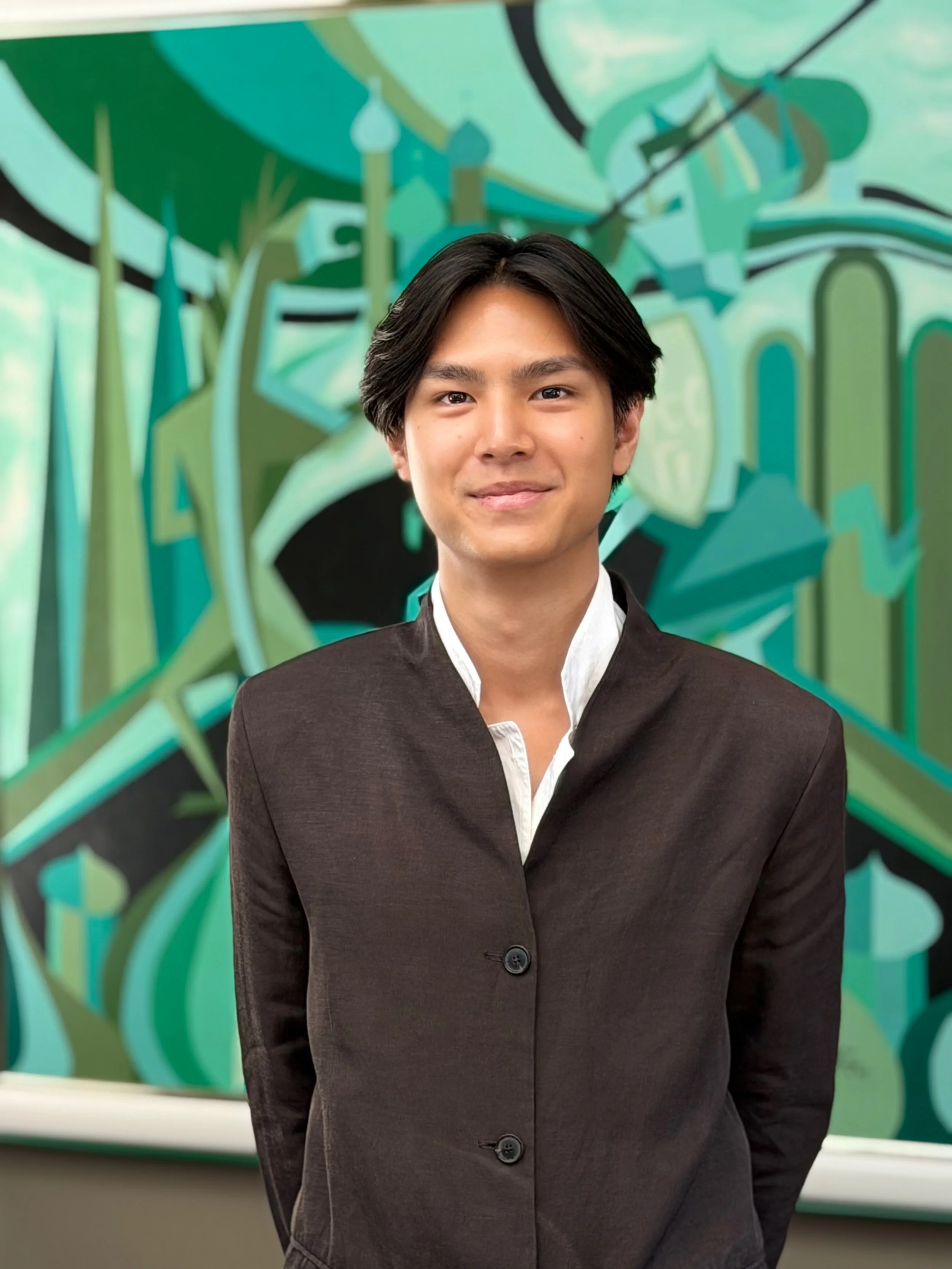 A young man with black hair and a slight smile, dressed in a black blazer with a white shirt underneath, standing in front of a colorful abstract mural with shades of green and blue.
