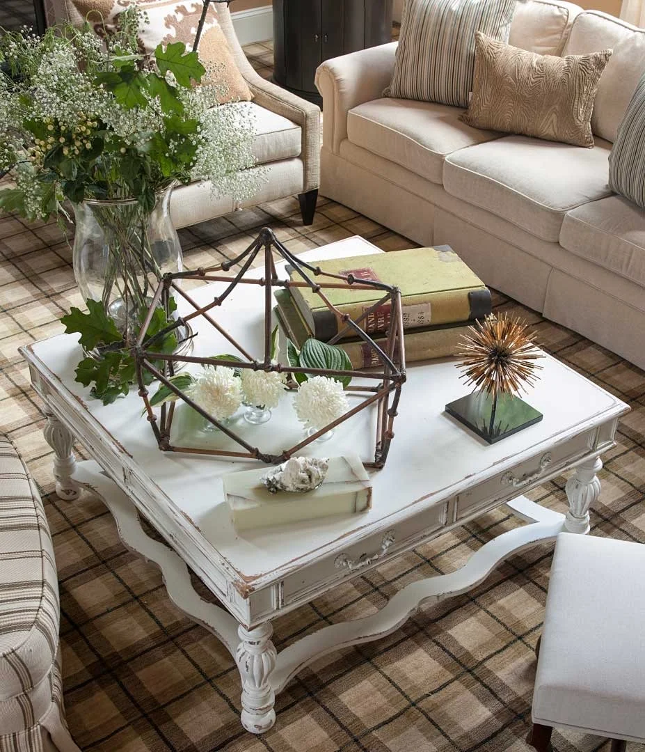 A living room coffee table with decorative objects including a large glass vase with greenery, a geometric tabletop sculpture, a gold spiked decorative object, and a couple of books. Surrounding the table are cream-colored sofas and chairs with strip