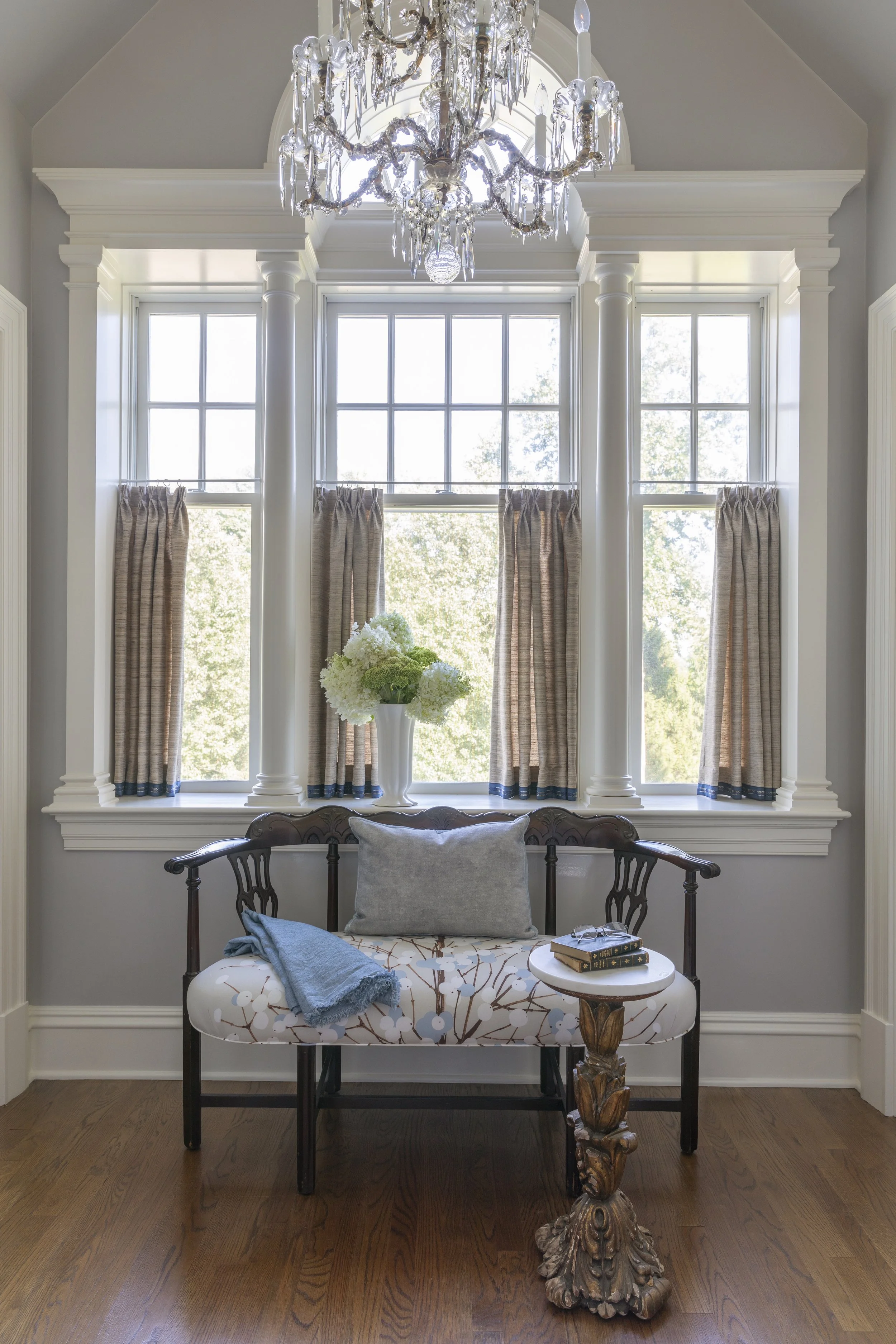 A living room with large windows, beige curtains, and white trim. An ornate chandelier hangs from the ceiling. In front, there is a patterned bench with black wooden legs, a gray pillow, a blue throw, and a small round side table with books and glass