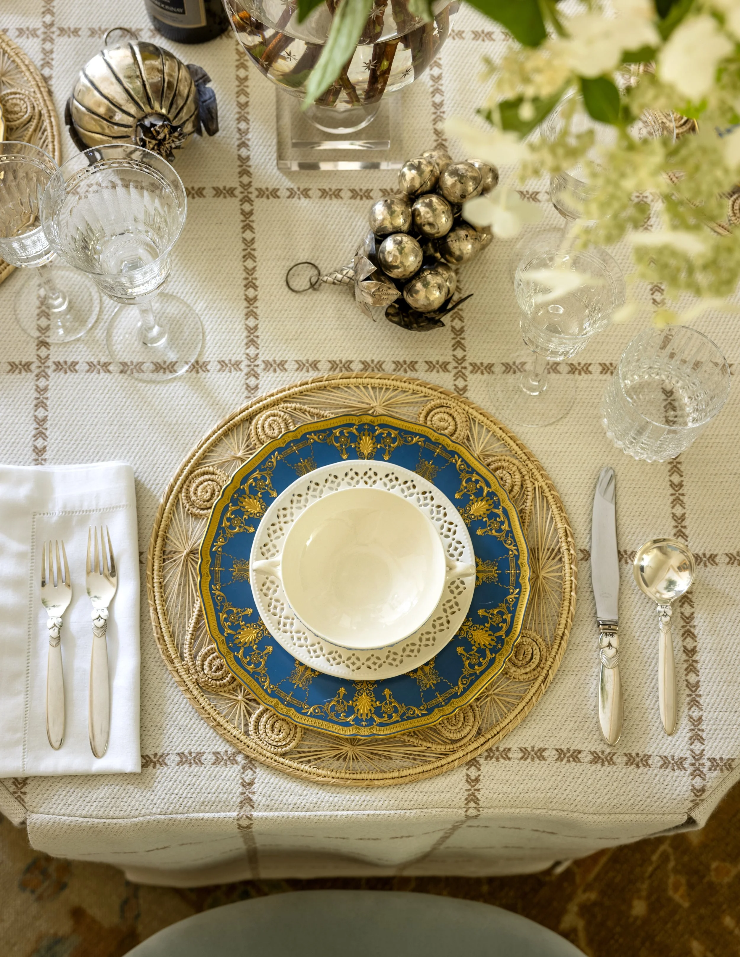 A formal table setting with a gold and blue patterned plate, white dinner plate, and intricate silverware on a white napkin. The table has a woven placemat, crystal glasses, a flower arrangement, and decorative silver ornaments, all on a tablecloth w