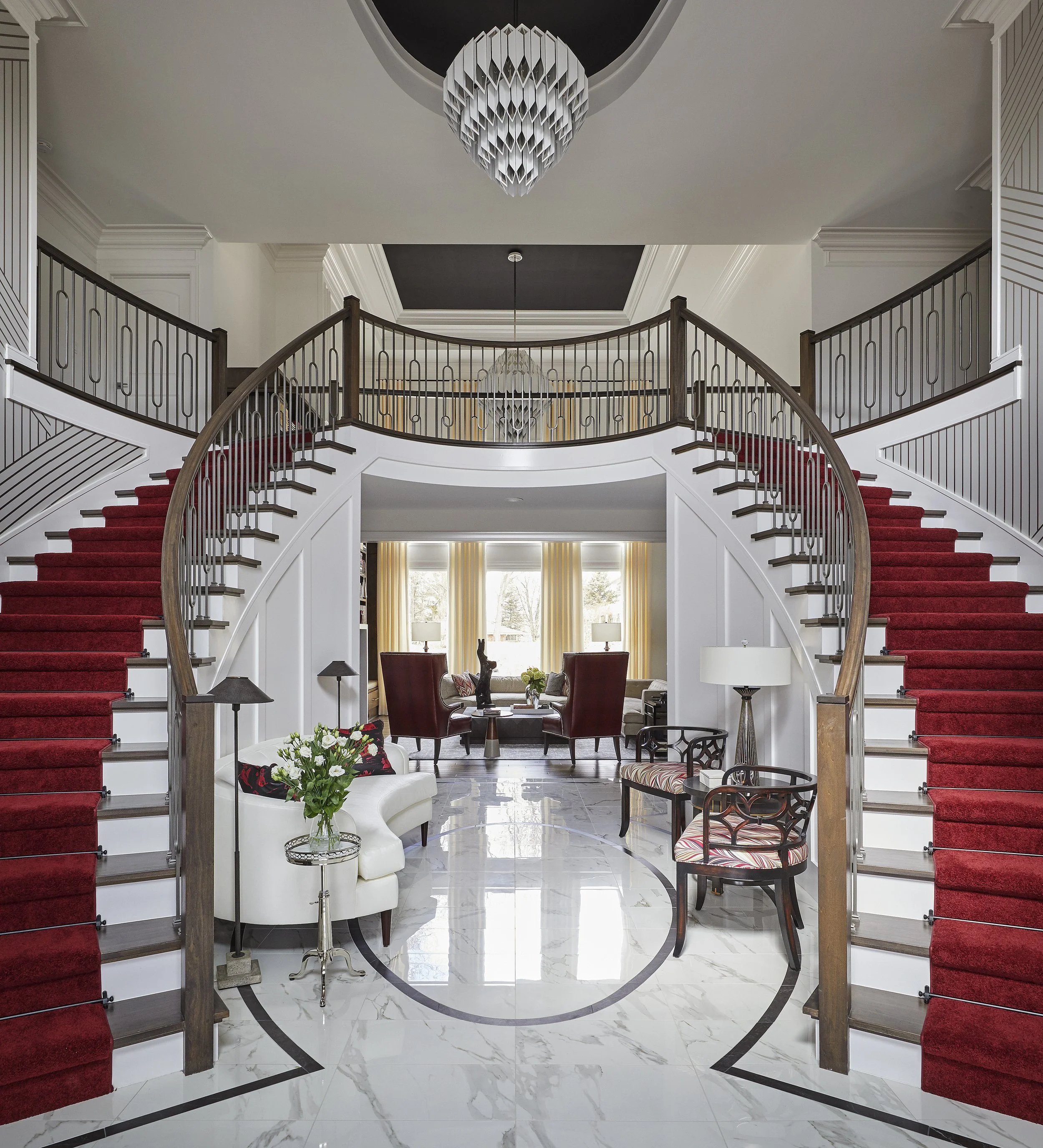 Elegant interior foyer with grand staircase covered in red carpet, white marble floor with black inlay, sitting area with white sofa, red chairs, and stone statues, large windows with yellow curtains, modern chandelier hanging from ceiling.