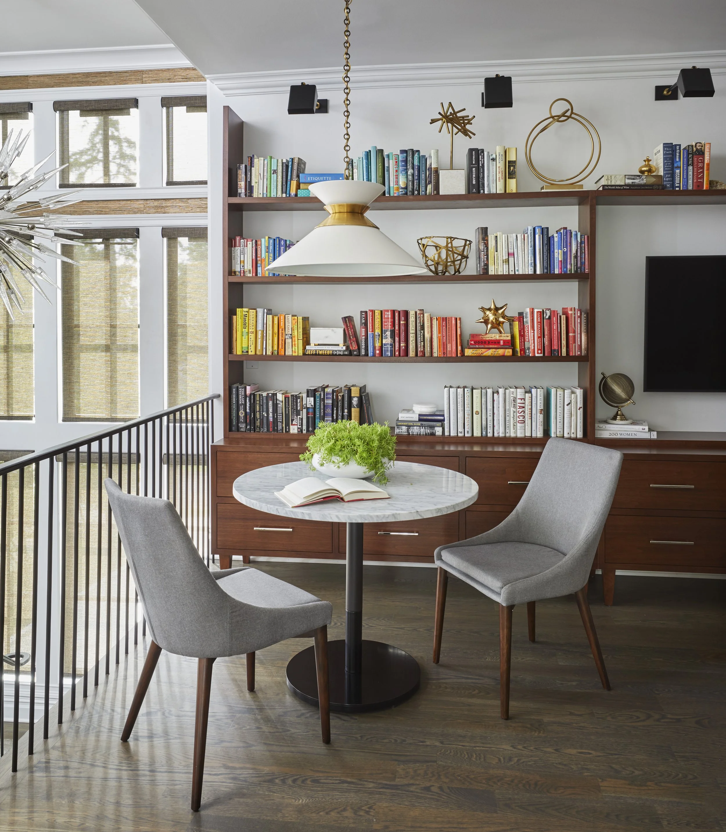 A corner of a living room with a bookshelf filled with colorful books, a round marble-top table with an open book and a green plant, two gray upholstered chairs, gold decorative objects, a flat-screen TV, and large windows with blinds letting in natu