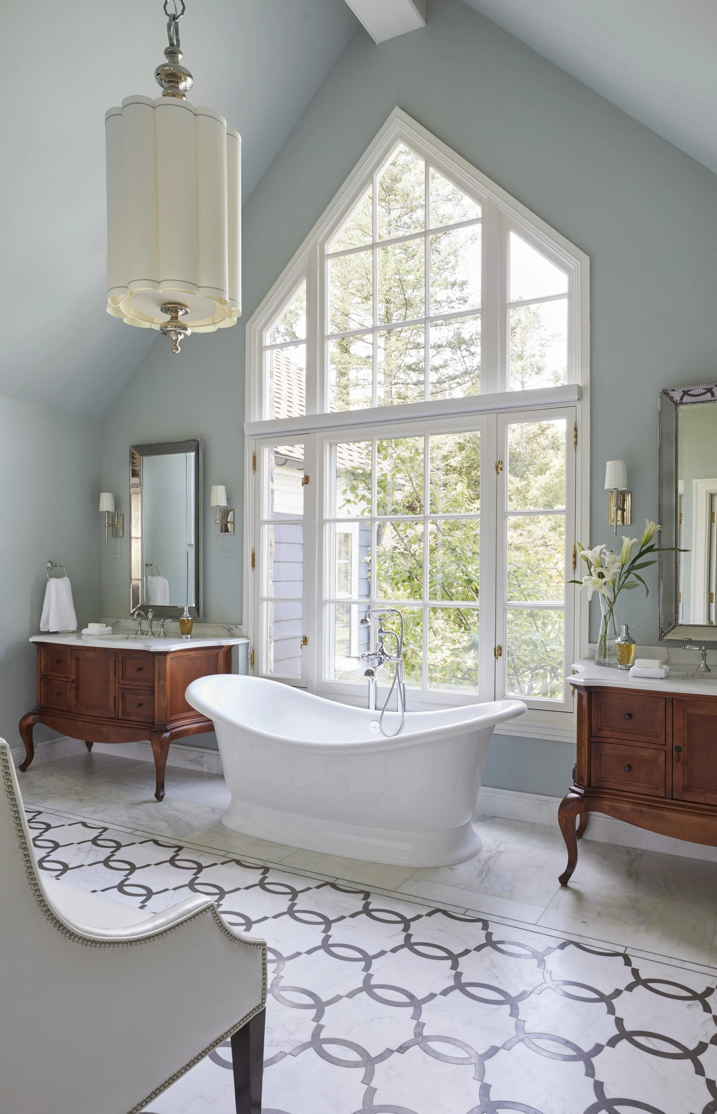 A bathroom with a freestanding white bathtub in front of a large, multi-pane window with a triangular top, overlooking trees outside. The bathroom features light blue walls, marble floors, vintage style wooden vanity cabinets, and wall-mounted mirror
