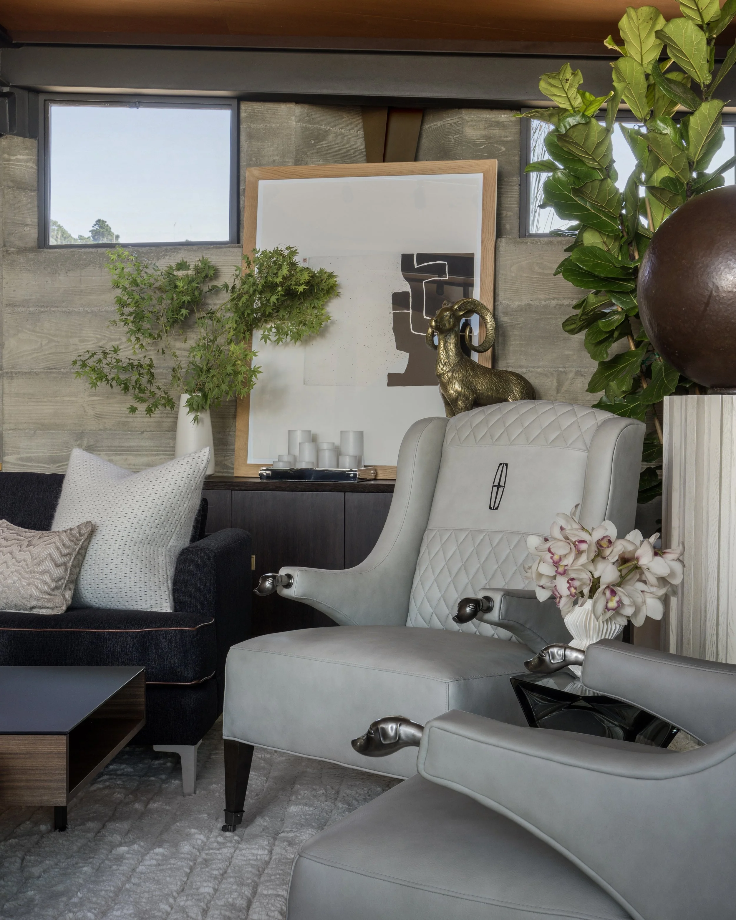 Living room with two windows, a large framed abstract art piece, a gold ram sculpture, a potted green plant, white upholstered chairs, a dark sofa with pillows, a wooden coffee table, a white orchid flower, and a gray rug.