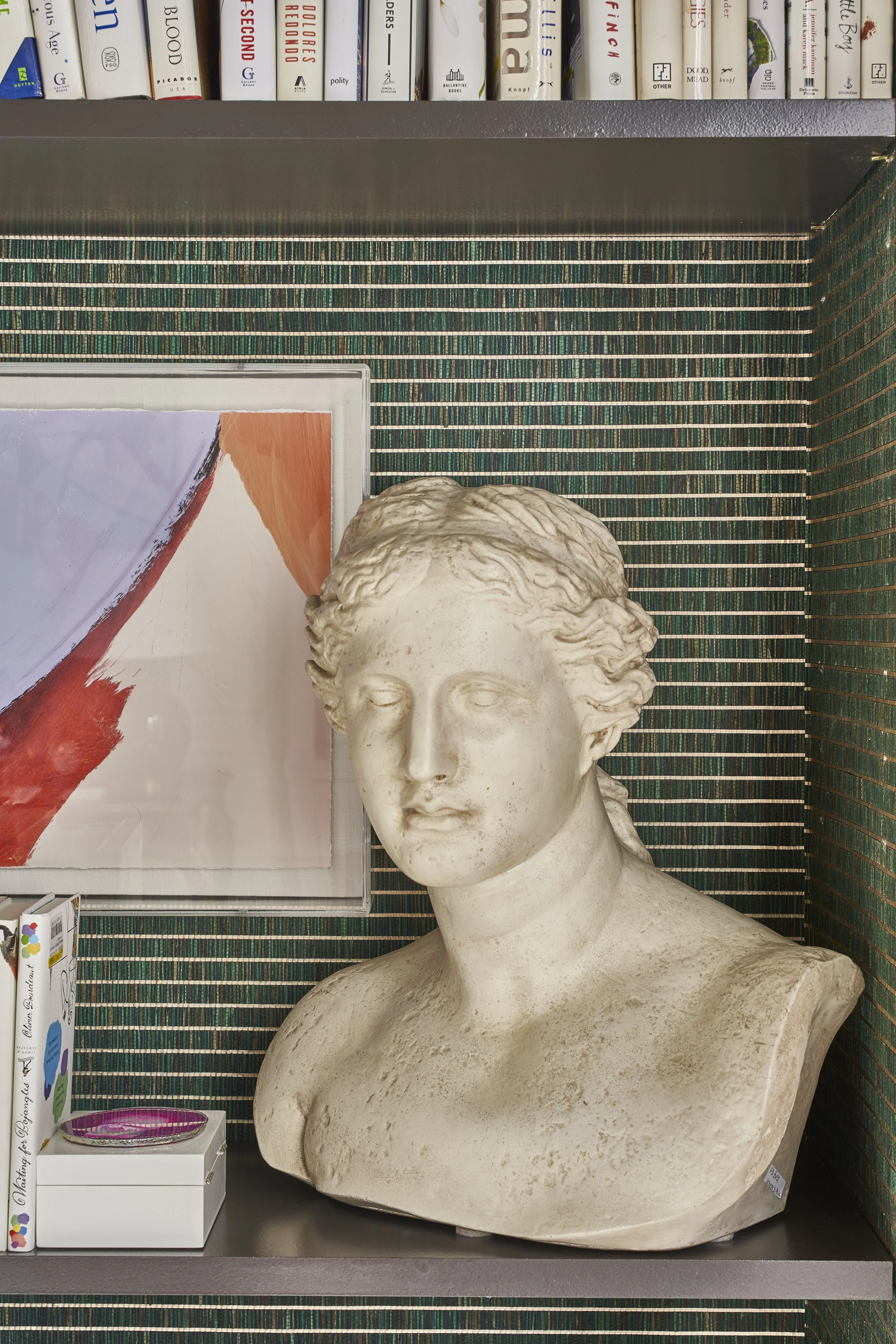A bookshelf with white books, a colorful abstract painting, a classical style bust sculpture of a woman with wavy hair, and a small decorative box on a gray surface. The background features green and black mosaic tiles.