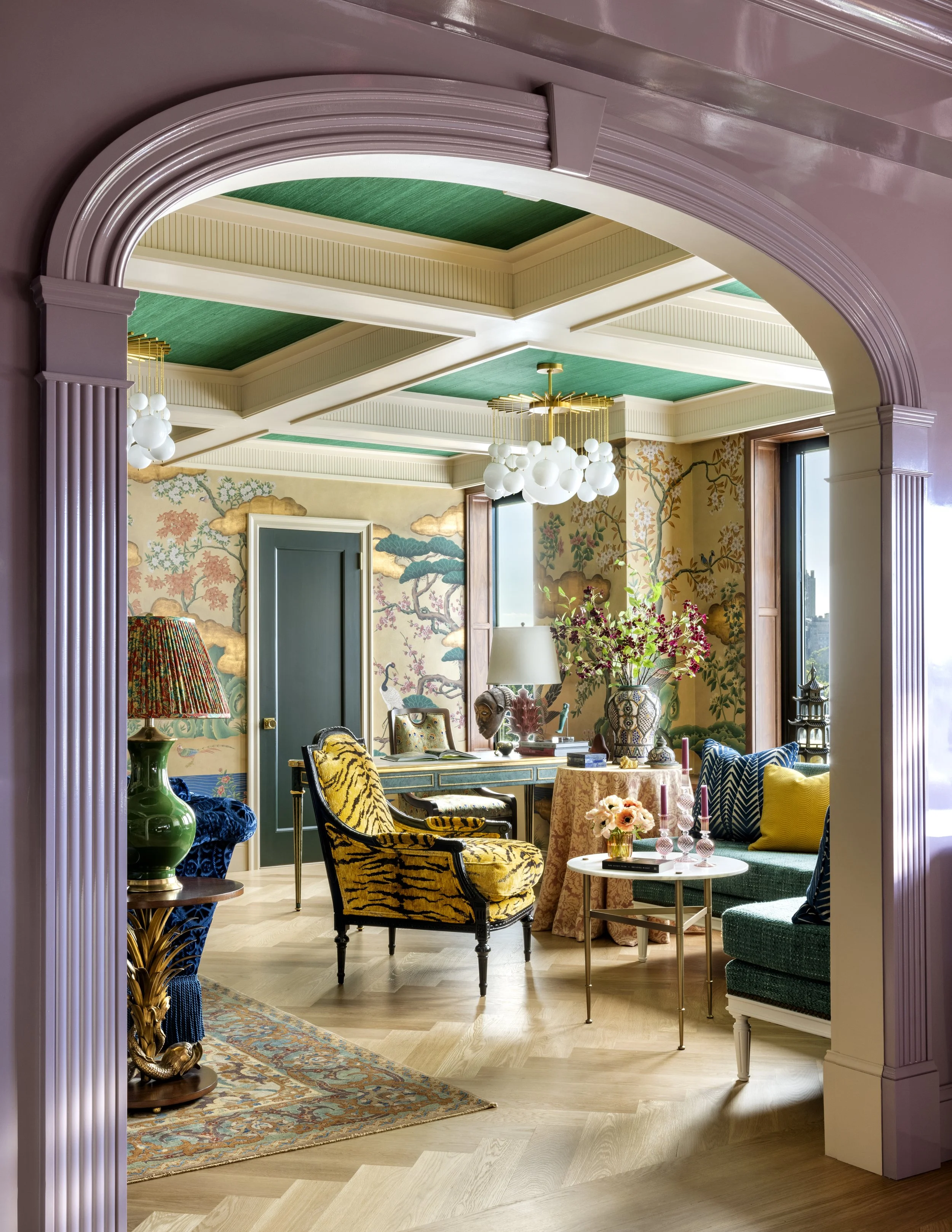 Interior of a colorful, eclectic living room viewed through an arched doorway with pink molding. The room features a patterned tapestry wall, a green sofa, vivid armchairs, a small round table with flowers, a vibrant rug, and decorative lighting fixt
