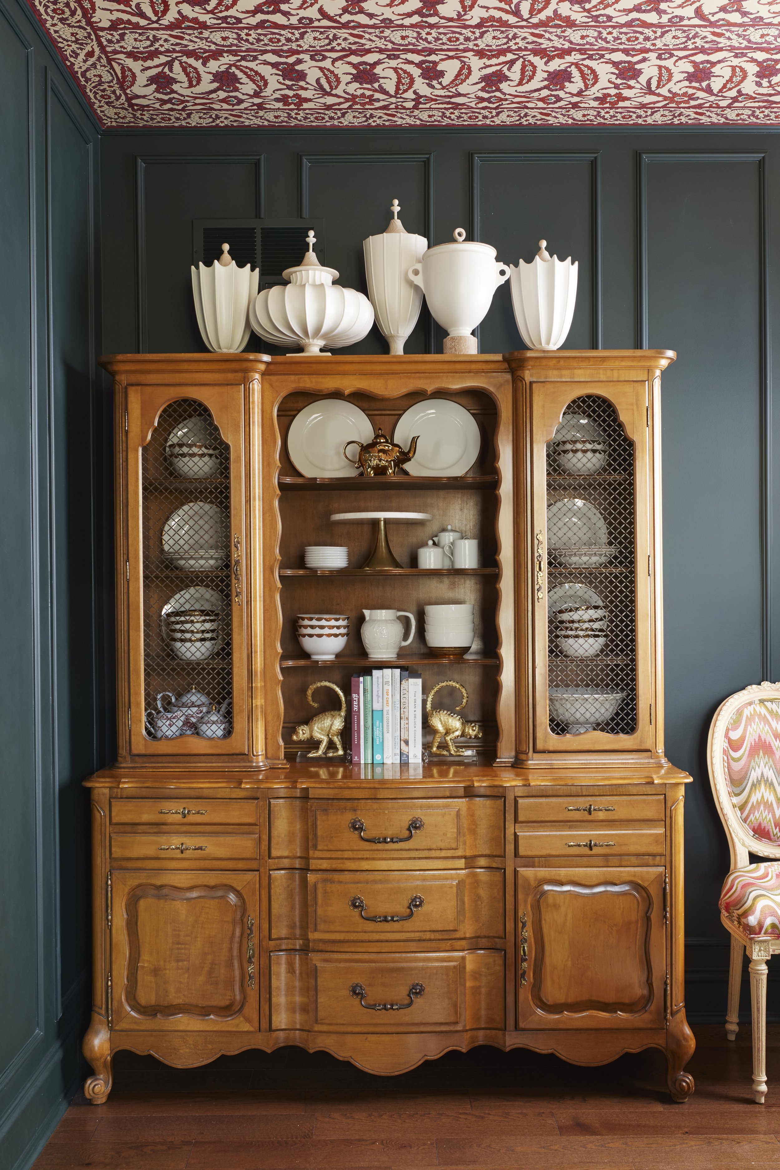 A wooden vintage china cabinet with glass-paneled doors, filled with white and patterned china, set against a dark teal wall with a decorative floral border on the ceiling. On top of the cabinet are white decorative vases, and inside the cabinet are 
