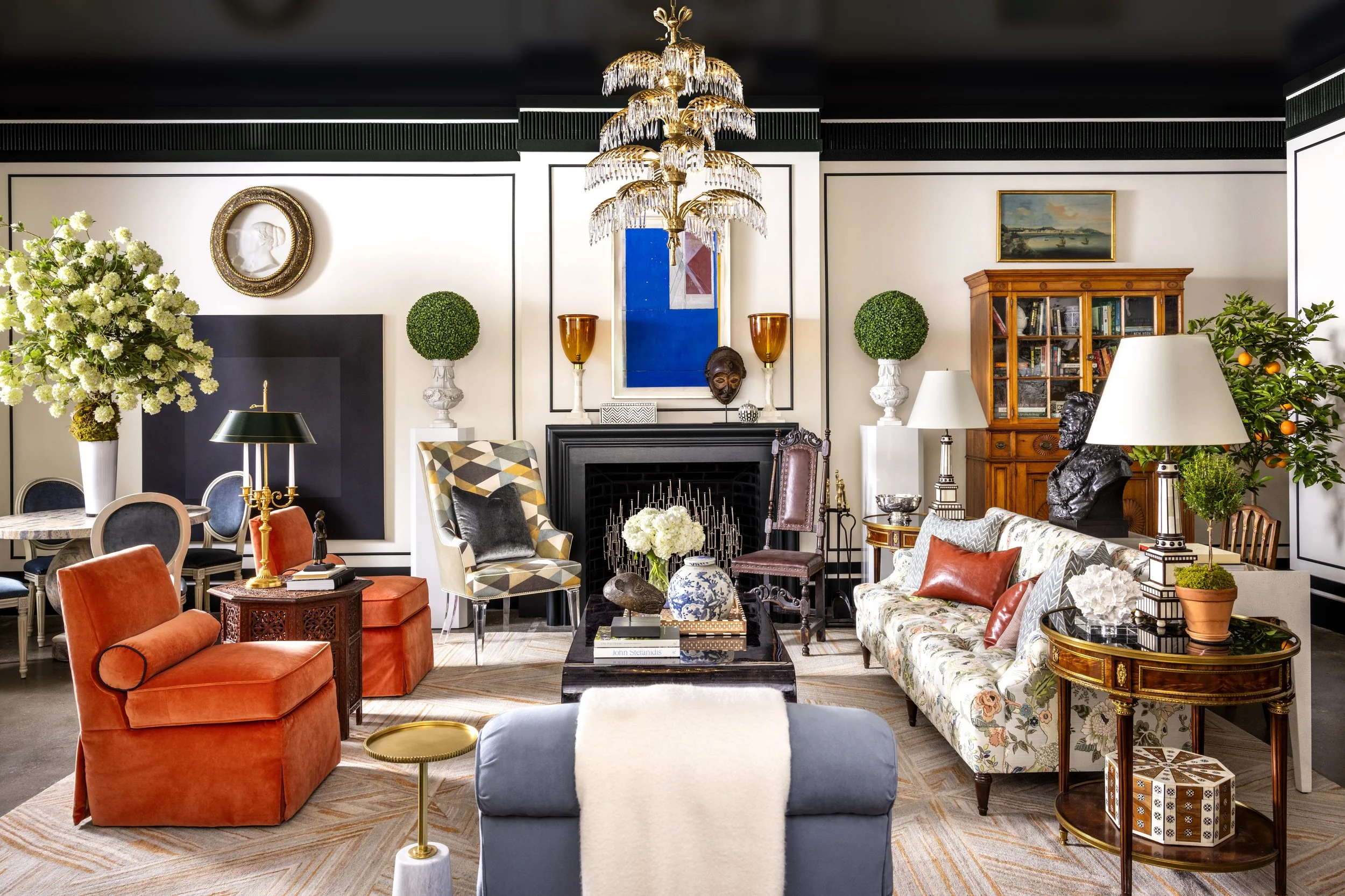 An elegant living room with a black fireplace, floral and abstract artwork, a gold chandelier, and a variety of colorful and patterned sofas and chairs. There are decorative plants, tables, and accessories adding to the stylish decor.
