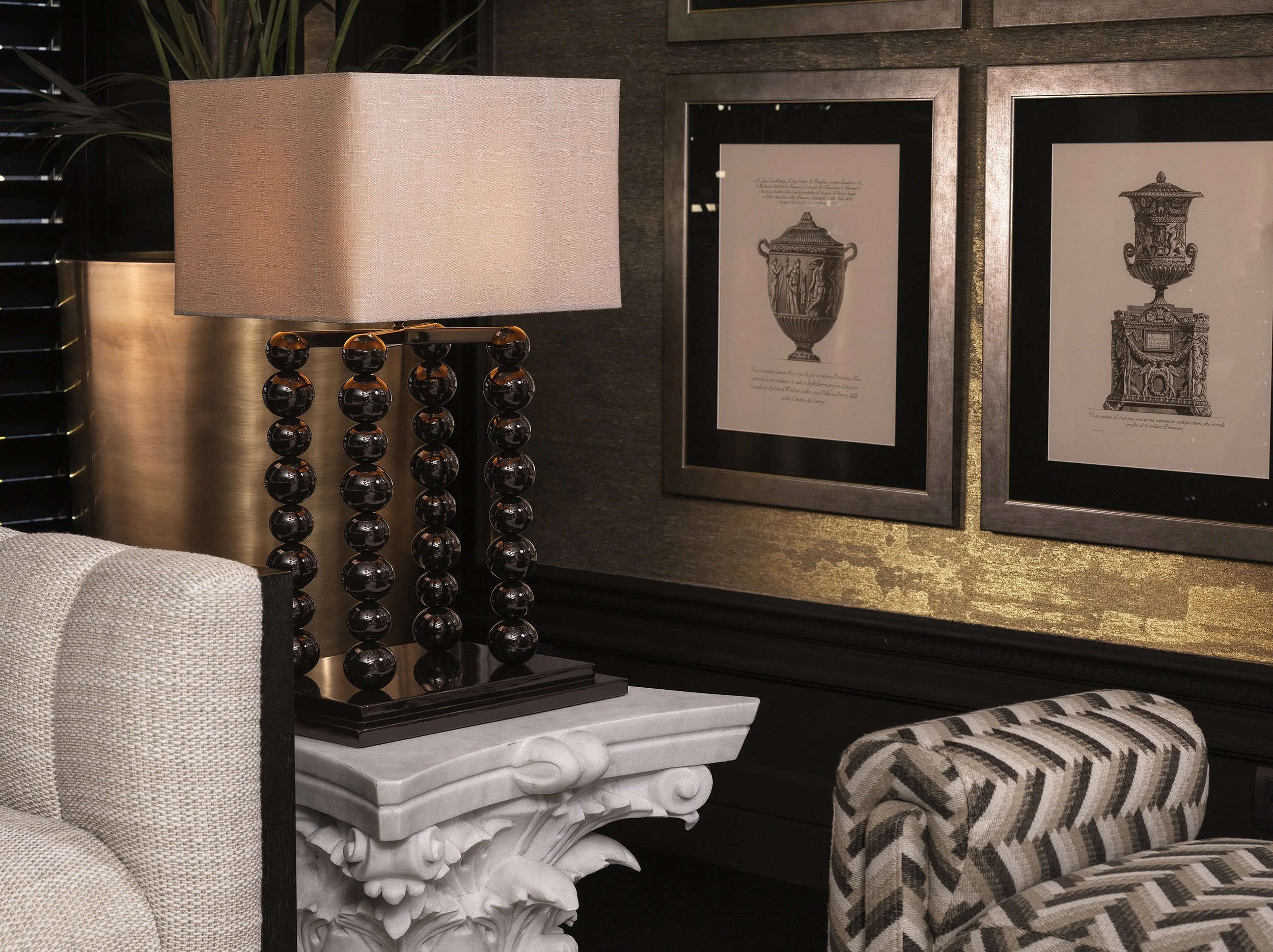 A table lamp with a beige fabric shade and a base made of black glossy spheres, standing on a white marble-like table with ornate detailing. Framed artwork featuring illustrations of decorative urns hangs on a dark textured wall behind the lamp, with