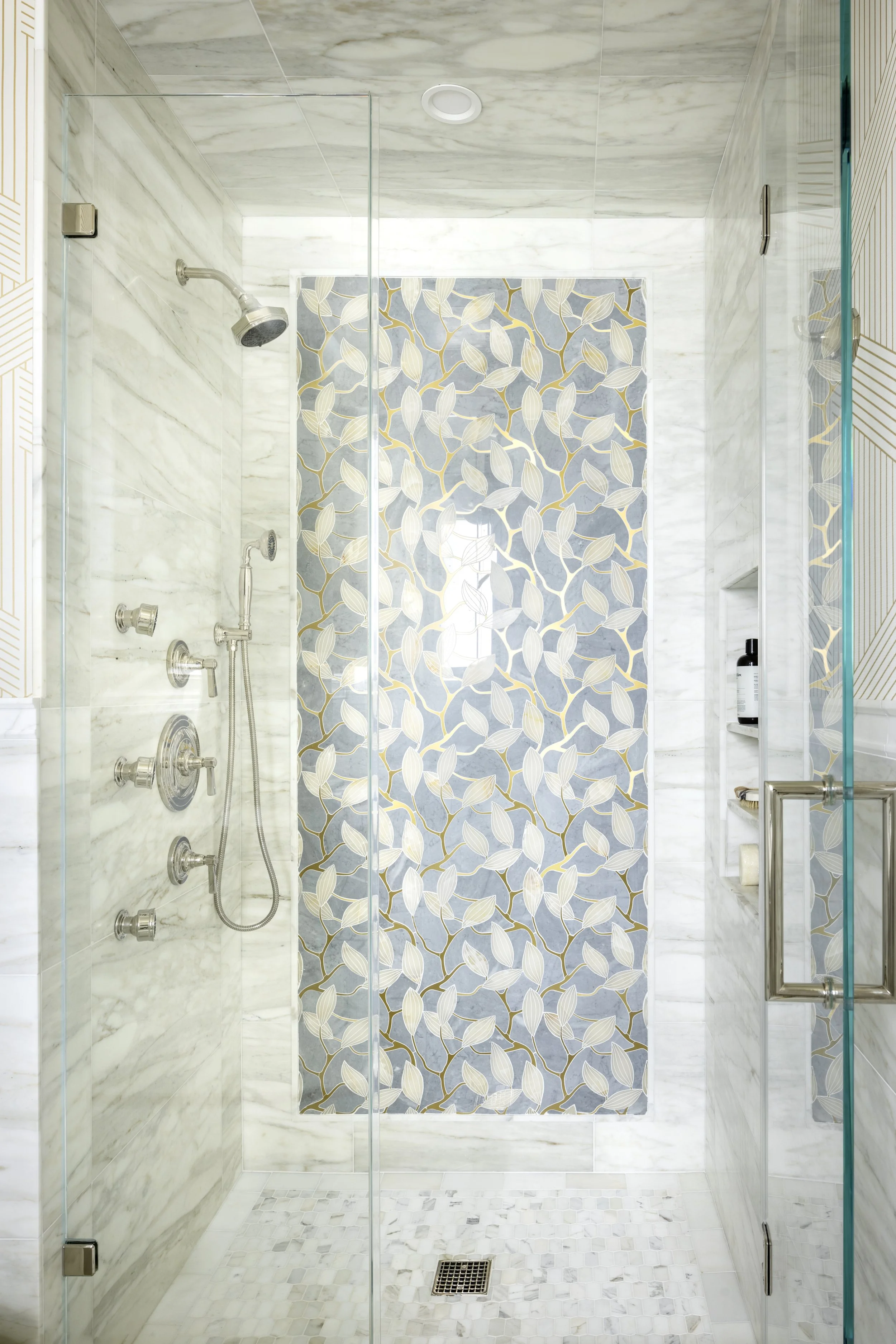 A walk-in shower with marble walls, a decorative tile panel with a leaf pattern, and a glass door.