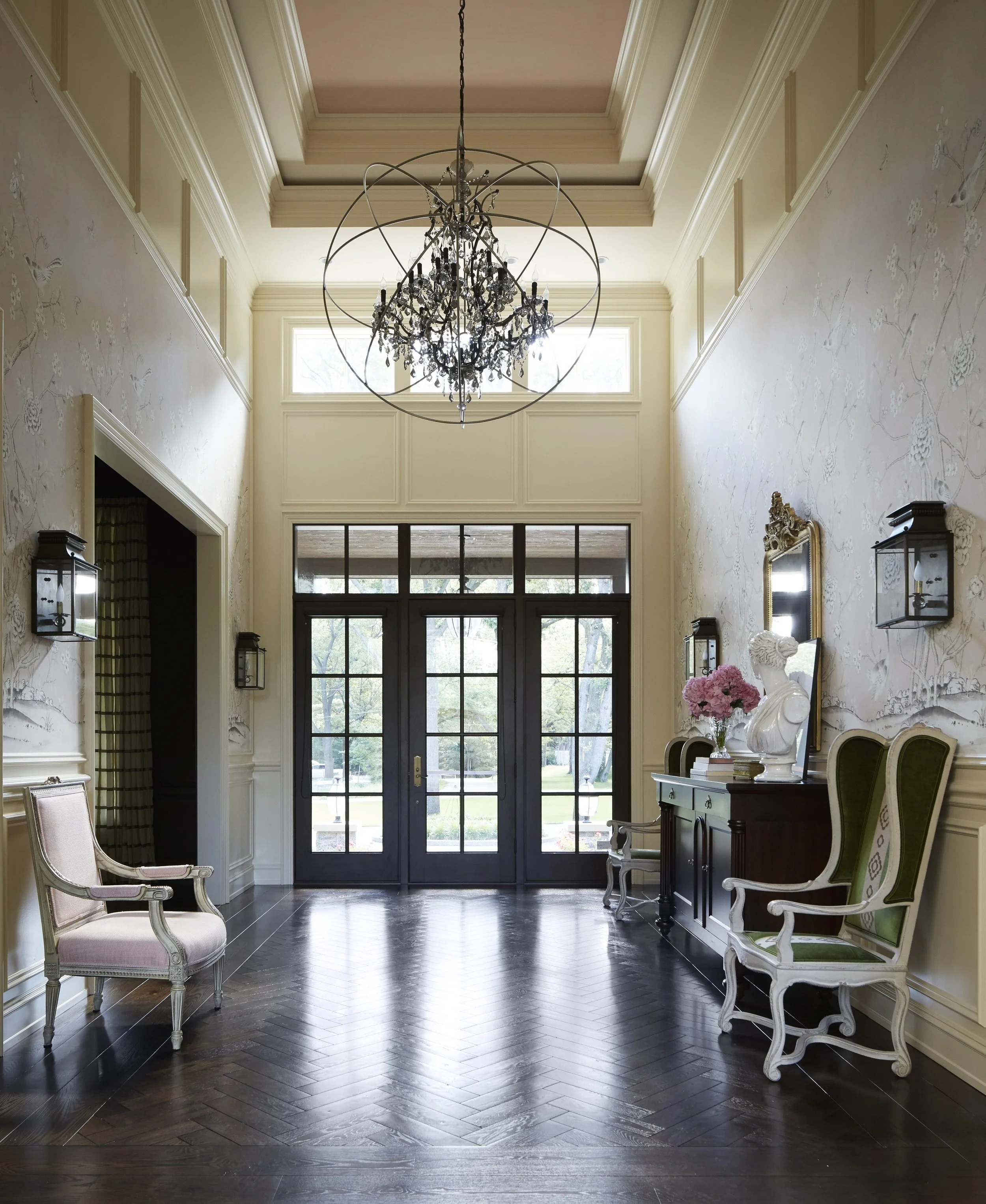 A spacious elegant foyer with a high ceiling, chandelier, large front door with multiple glass panes, and a mix of vintage and modern furniture, including pink and green upholstered chairs, a vase of pink flowers, and decorative wall sconces.