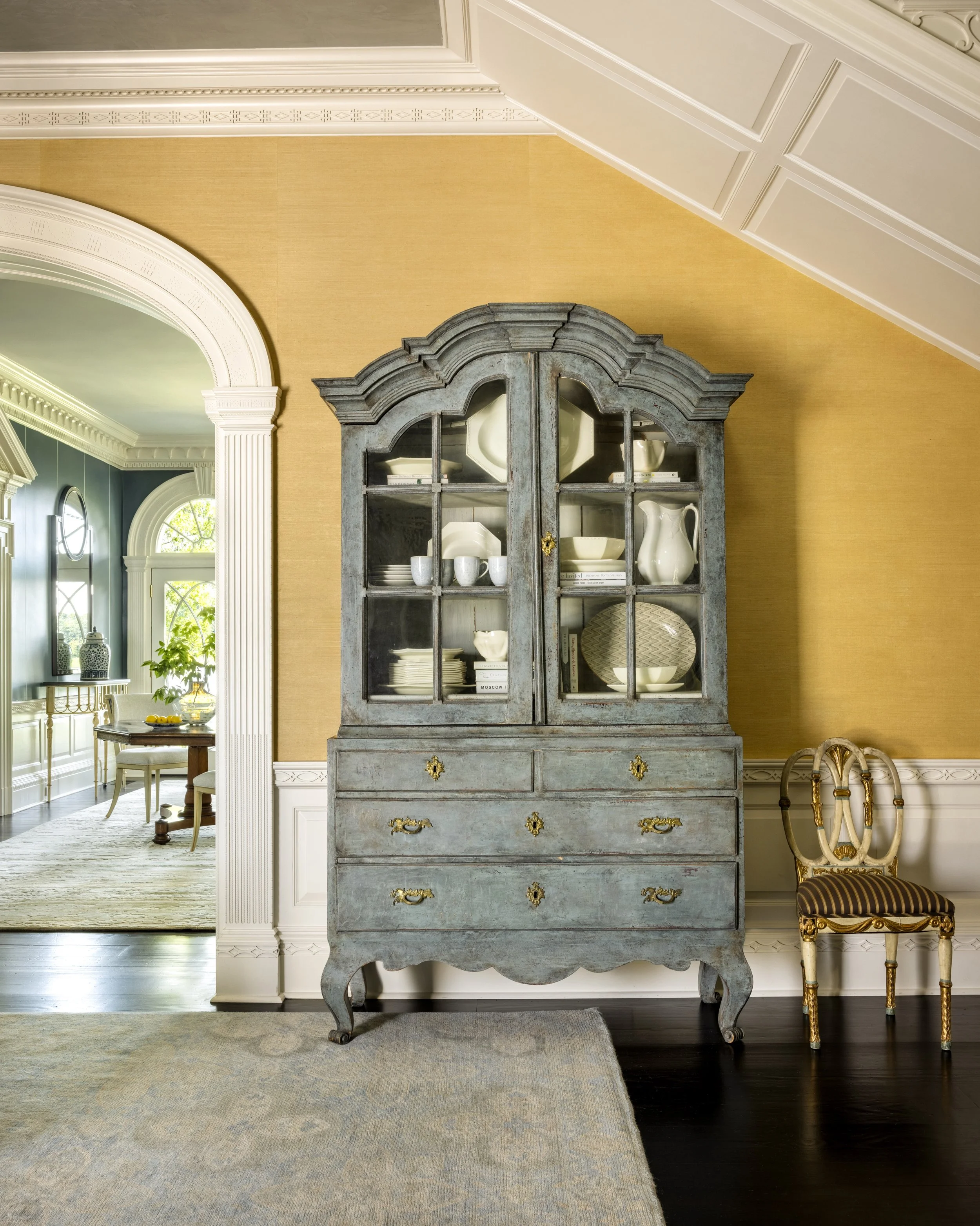 A vintage blue-gray wooden cabinet with glass doors displaying white dishware and books, set against a gold-colored wall in a traditionally decorated dining room. To the right, a decorative wooden chair with gold accents and striped upholstery is vis