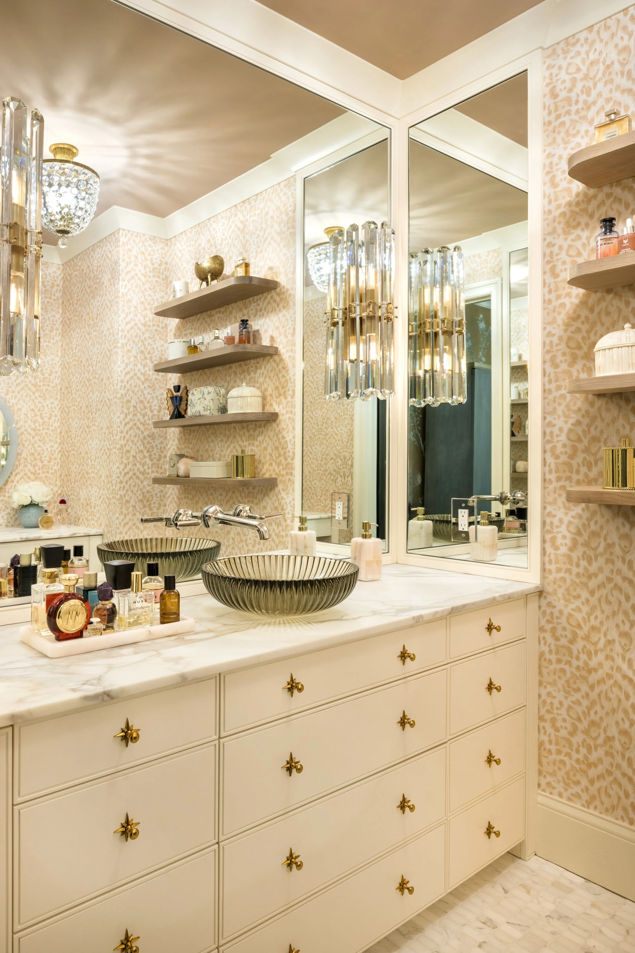 A bathroom vanity with two round black and gold sinks, a marble countertop, and a large mirror with a white frame. The wall behind the vanity is decorated with peach leopard print wallpaper, and there are floating shelves with various toiletries and 