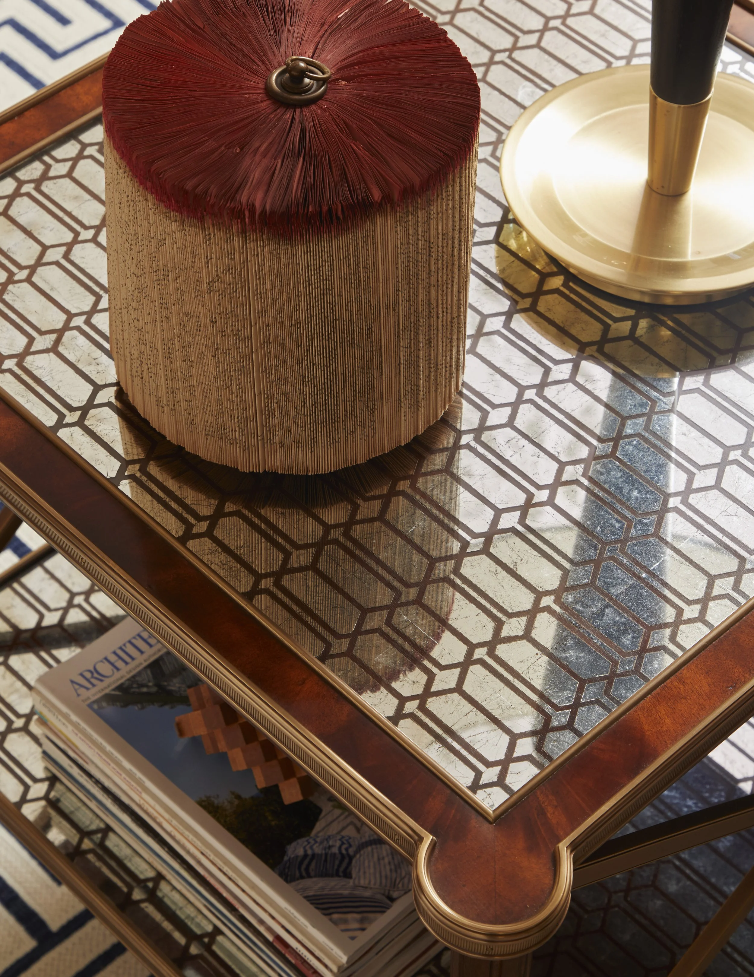 A glass-top table with a geometric-patterned surface, featuring a decorative item and a stack of magazines underneath.