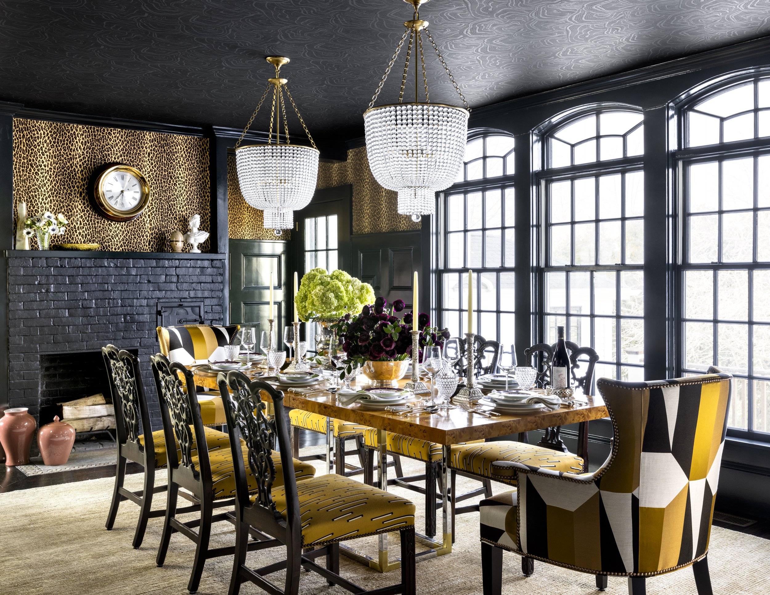 A dining room with a large wooden table, several black and yellow chairs, and a floral centerpiece. The room has black walls with white trim, patterned wallpaper, black brick fireplace, large window with black framing, and 2 chandeliers hanging from 