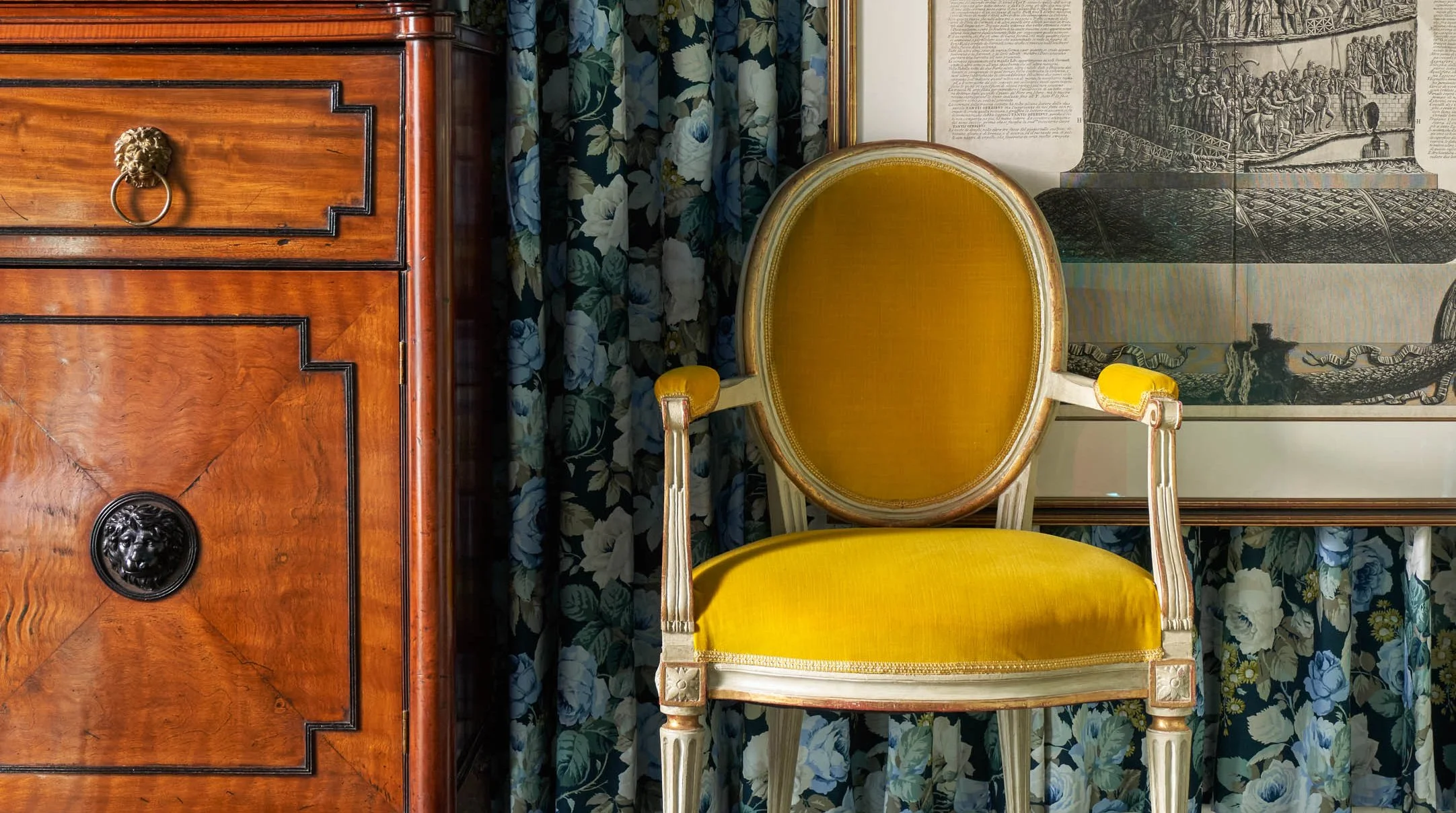 An antique wooden desk with lion head drawer pulls, next to a mustard yellow upholstered chair with ornate details. Behind the chair, there is a framed tapestry or wall hanging depicting historical or mythological scenes, and a floral curtains in blu