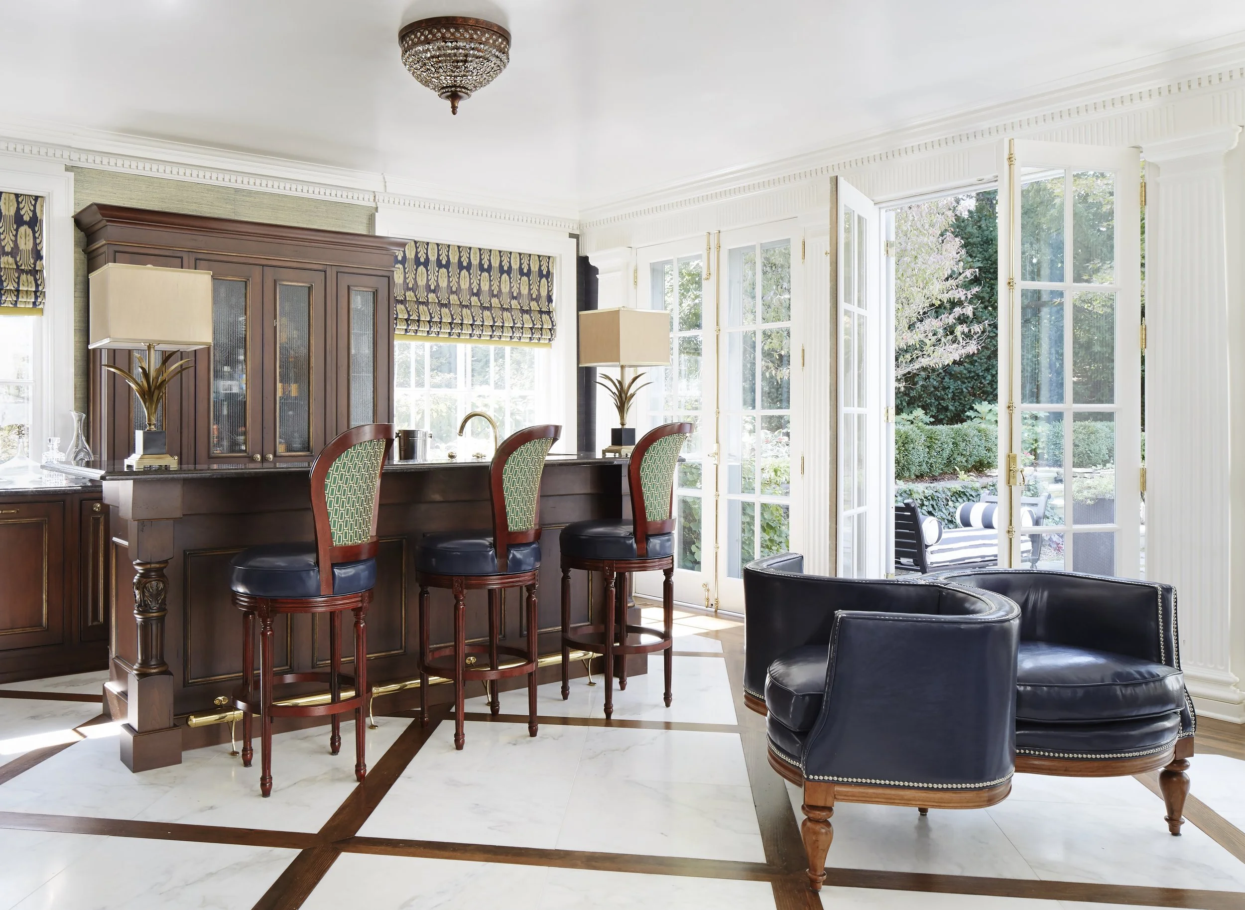 A bright living room with large windows, a dark wooden bar counter with green patterned chairs, a black leather armchair, and a ceiling chandelier.