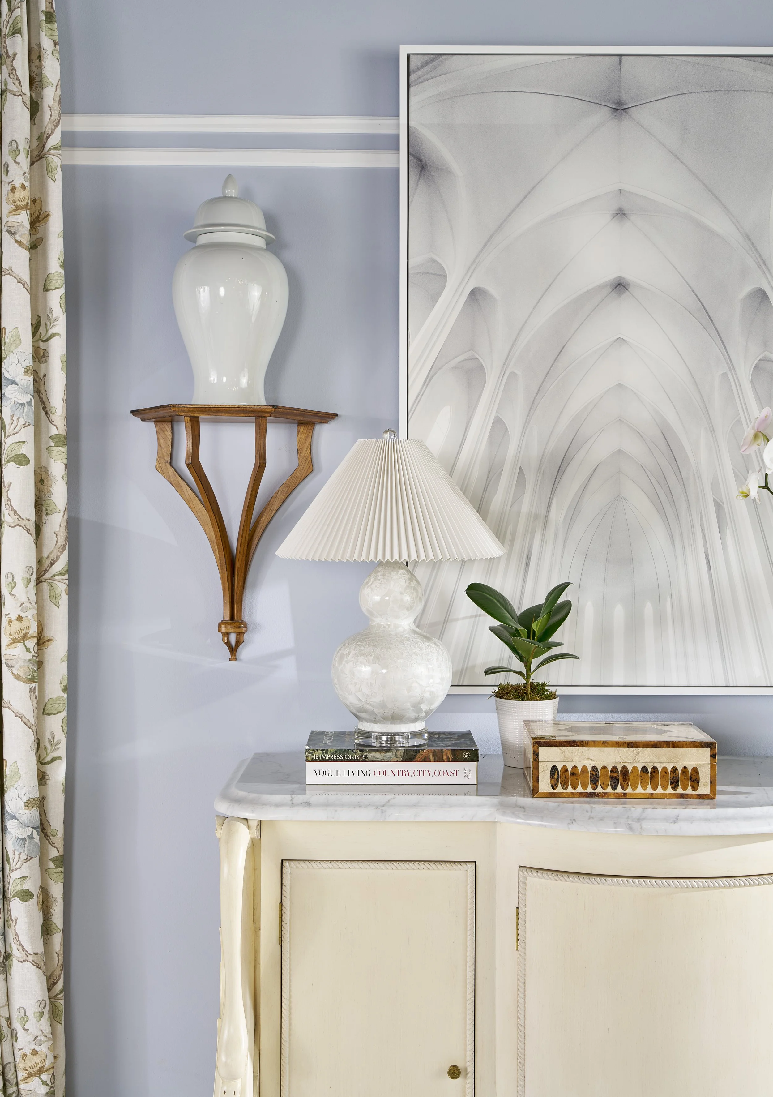 Interior decor with a white lamp, a small potted plant, a large abstract artwork showing vaulted brick ceilings, and a marble table with books and decorative objects. A wooden wall shelf holds a white vase, and floral curtains frame the left side.