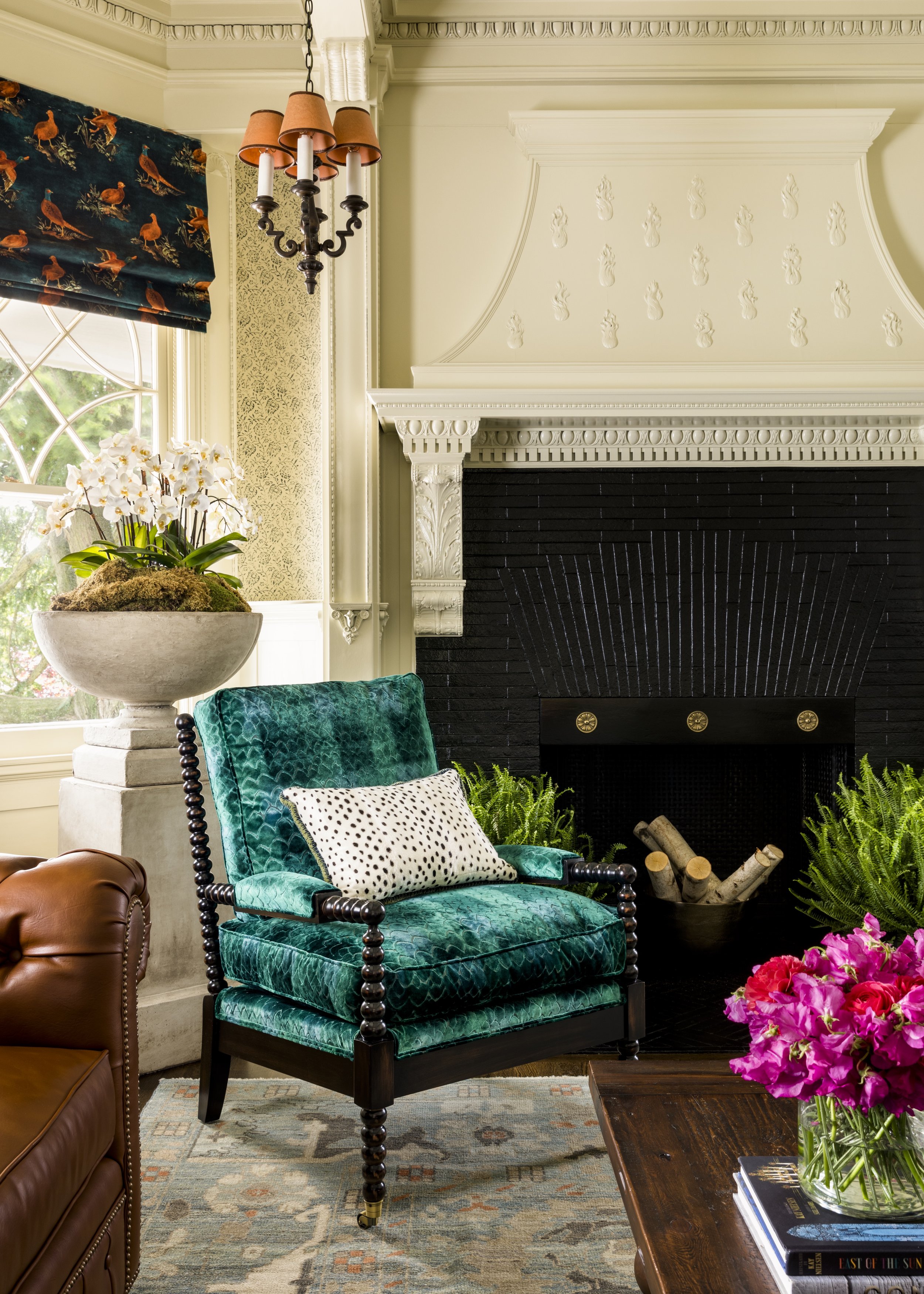 Interior living room with a large black fireplace, teal velvet armchair with a white polka dot pillow, brown leather chair, potted orchid, floral rug, window with a duck-patterned curtain, chandelier, and pink flowers on a wooden table.
