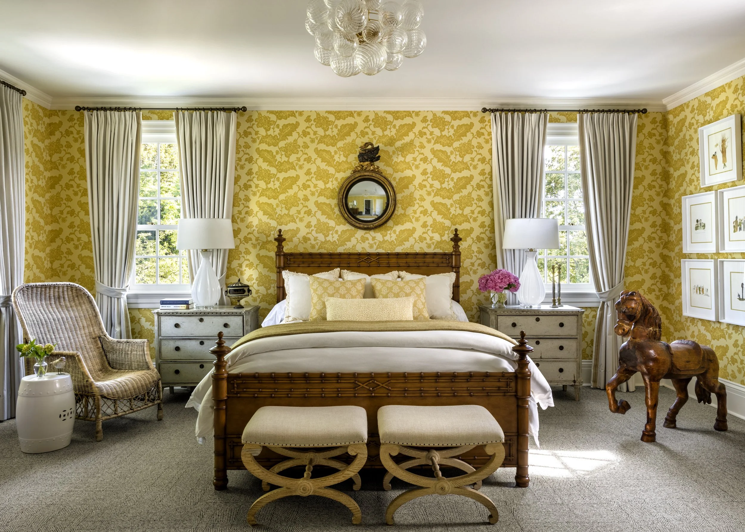 A bedroom with yellow floral wallpaper, a wooden bed with cream and yellow bedding, white side tables, white lamps, and a mirror on the wall. There are two windows with white curtains and framed artwork on the wall, and a decorative wooden horse scul