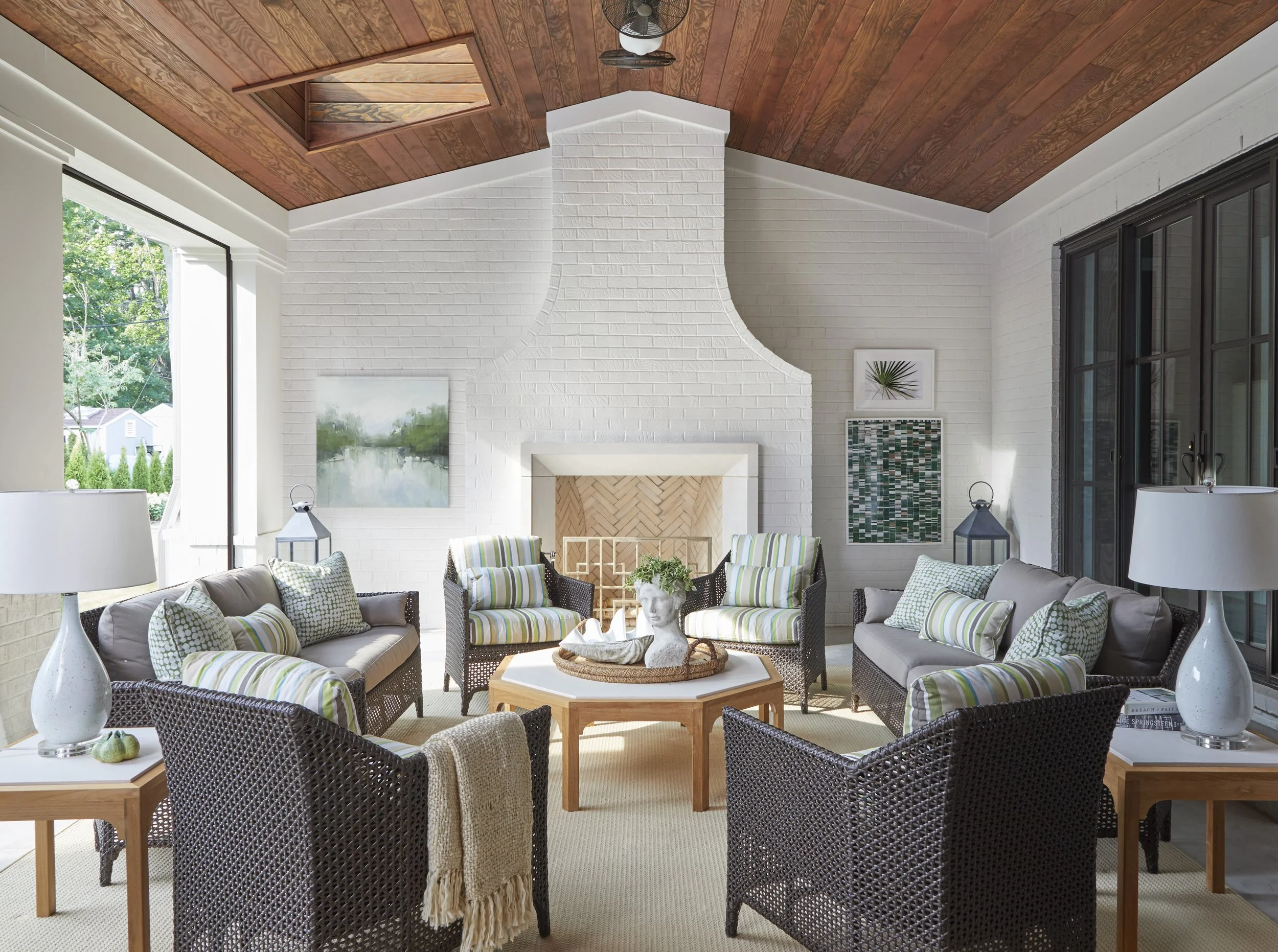 A bright living room with white brick walls, a vaulted wood ceiling, large windows, and assorted furniture including wicker chairs with green and white striped cushions, tables with lamps, and wall art.