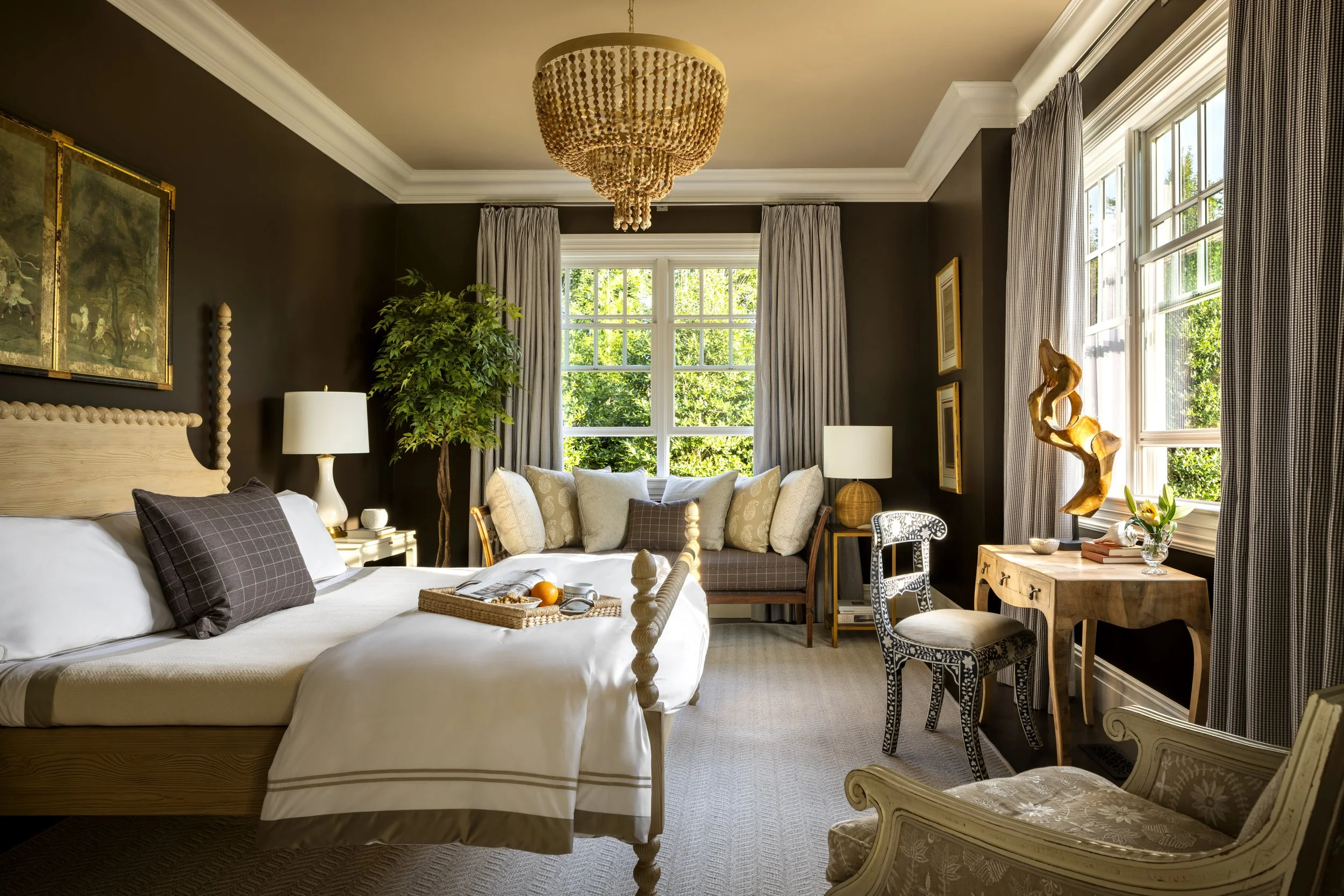 A bedroom with a large bed with white bedding and patterned pillows, a wooden headboard, a tray with breakfast on the bed, a seating area with a cushioned sofa, a side table with a lamp, a tree inside the room, large windows with curtains, a chandeli