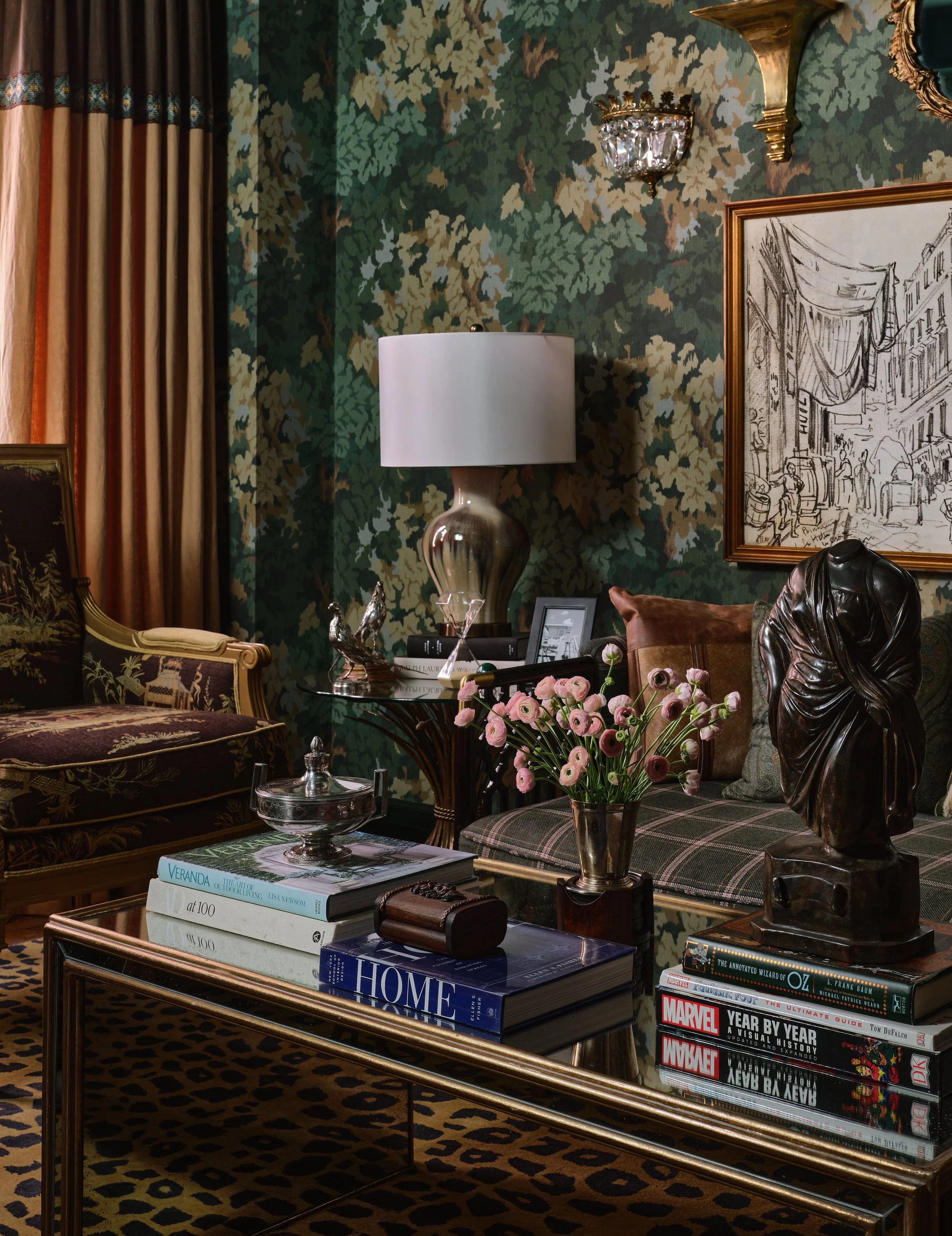 An elegant, vintage-style living room with floral wallpaper, a floral couch, a glass coffee table with books, a white lamp, a decorative sculpture, pink flowers, framed artwork, and a leopard print rug.