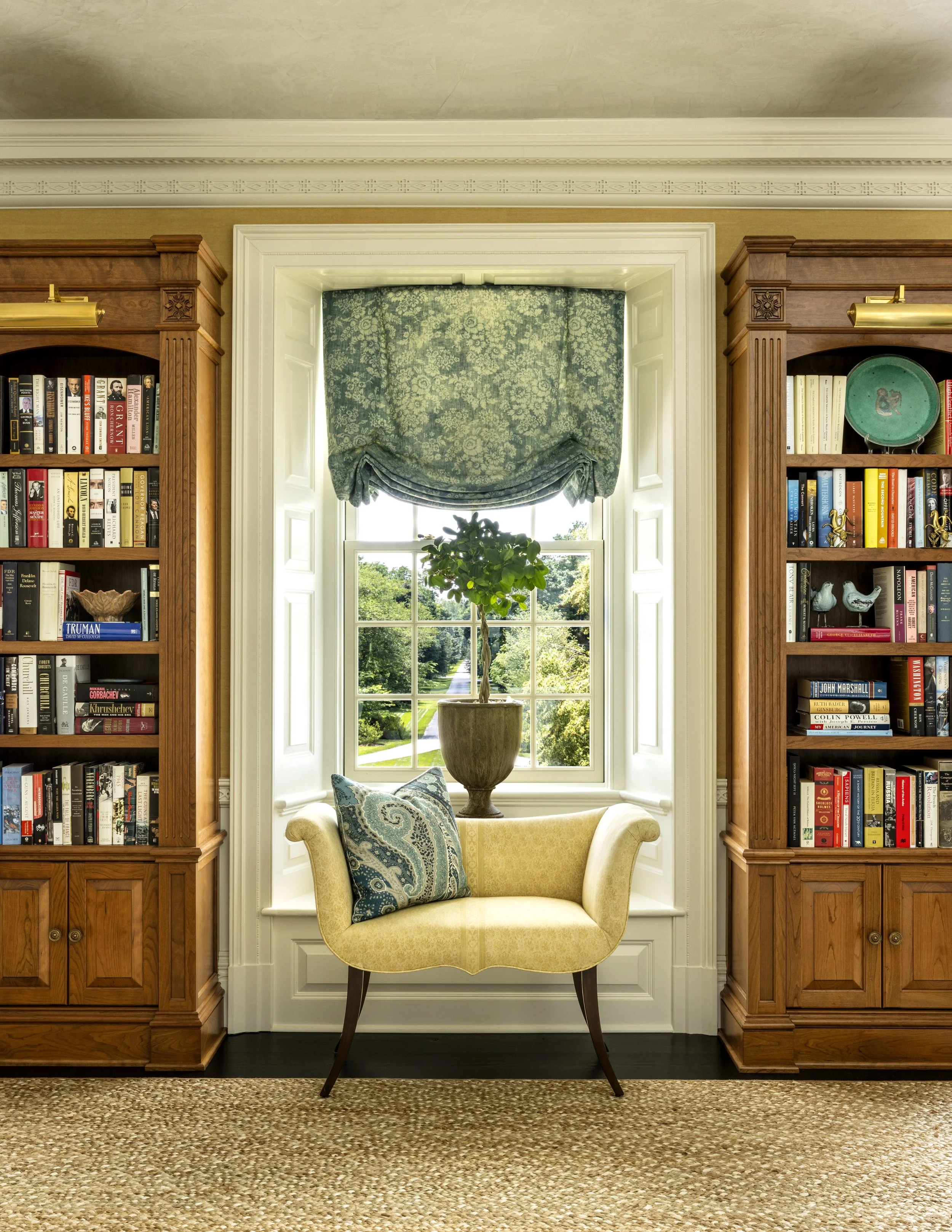 A cozy reading nook with a yellow upholstered armchair, a decorative pillow, and a window with a decorative green curtain. There are bookshelves on each side of the window filled with books and decorative items, and a small plant on the windowsill.