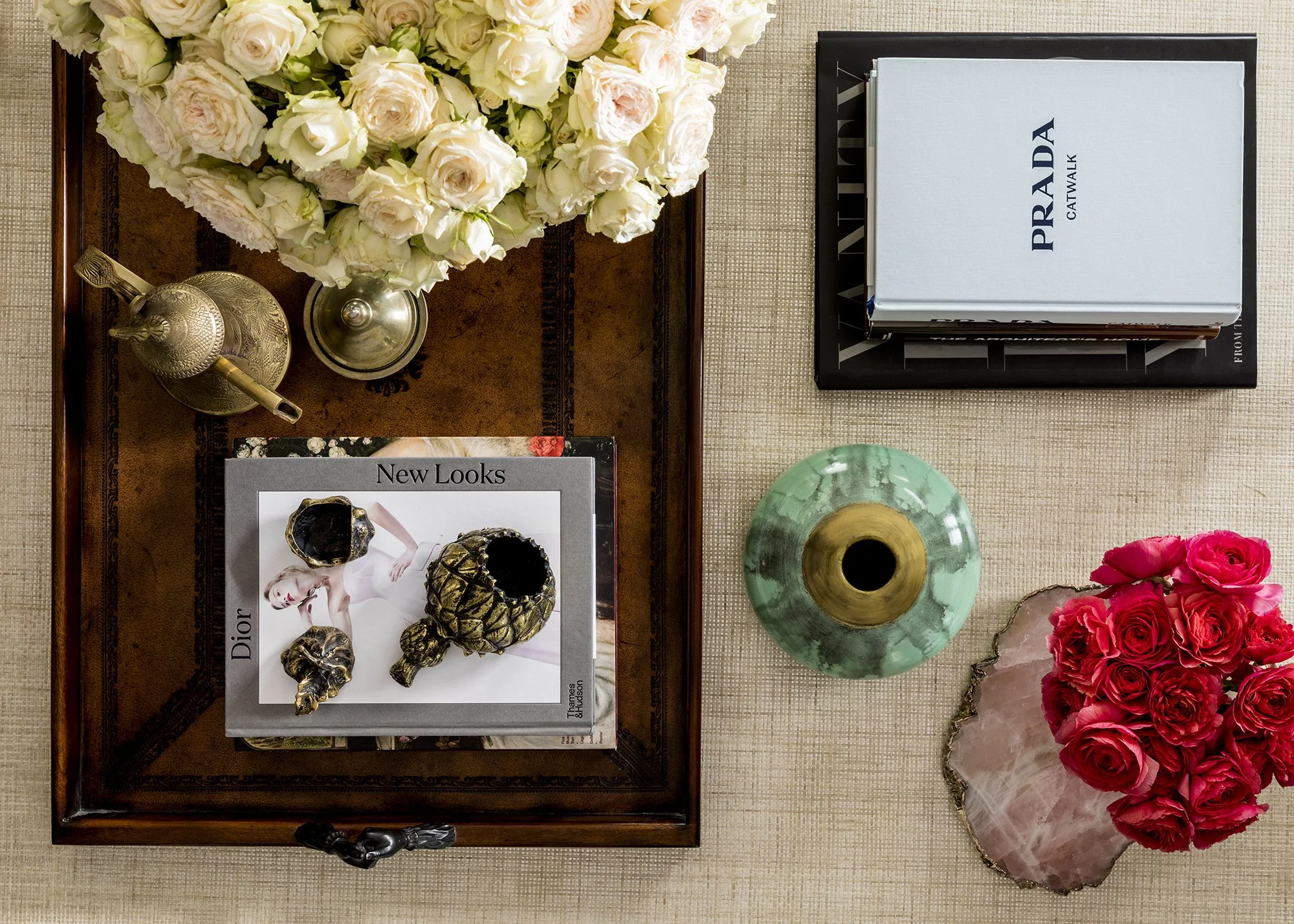 A decorative display with a bouquet of white and pale pink roses, gold and silver decorative objects, a Prada Catwalk book, a Dior magazine, a green marble vase, and pink roses on a beige fabric surface.