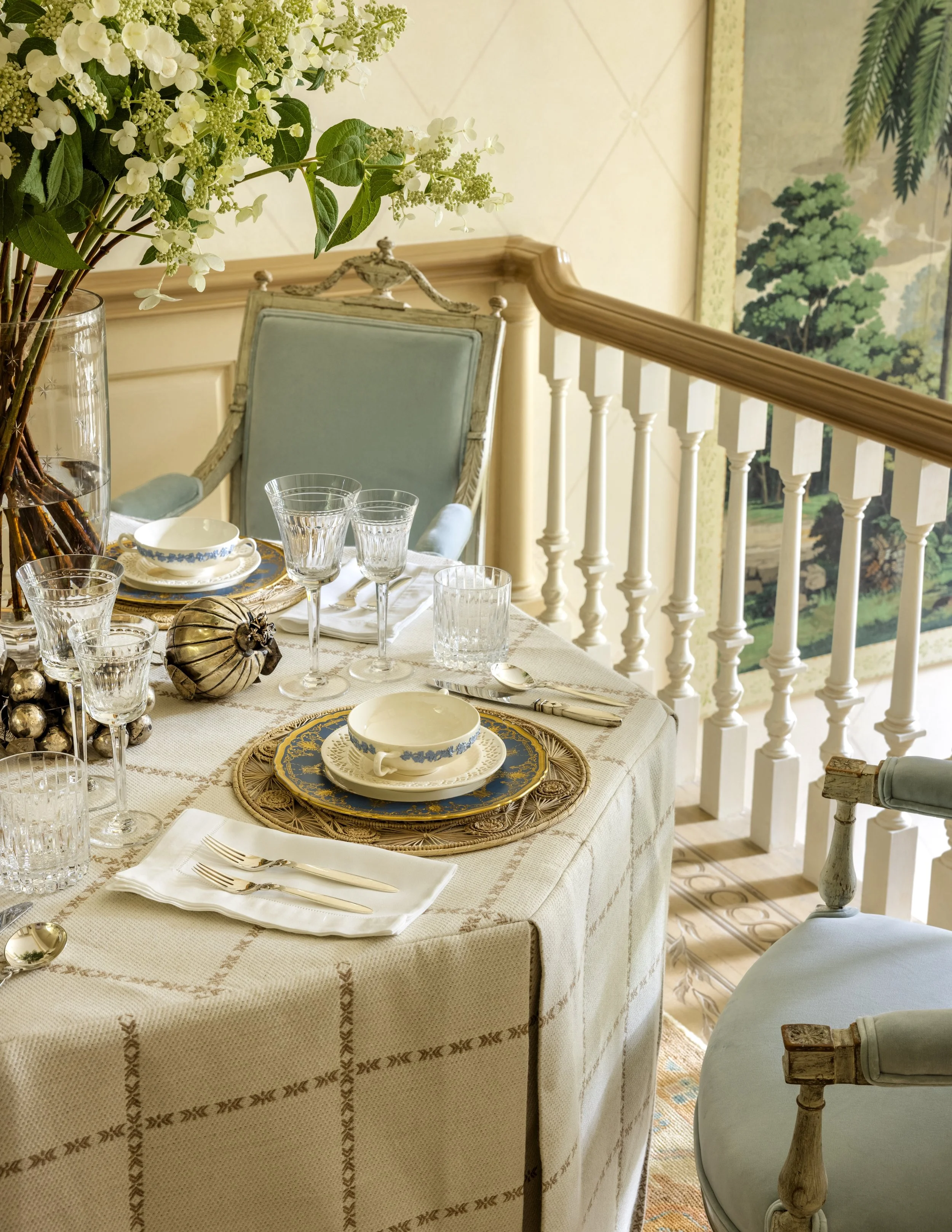 A formal dining table set with fine china, crystal glasses, silverware, and decorative placemats. The table is covered with a cream-colored tablecloth featuring embroidered patterns, with a centerpiece of white flowers in a glass vase. The background