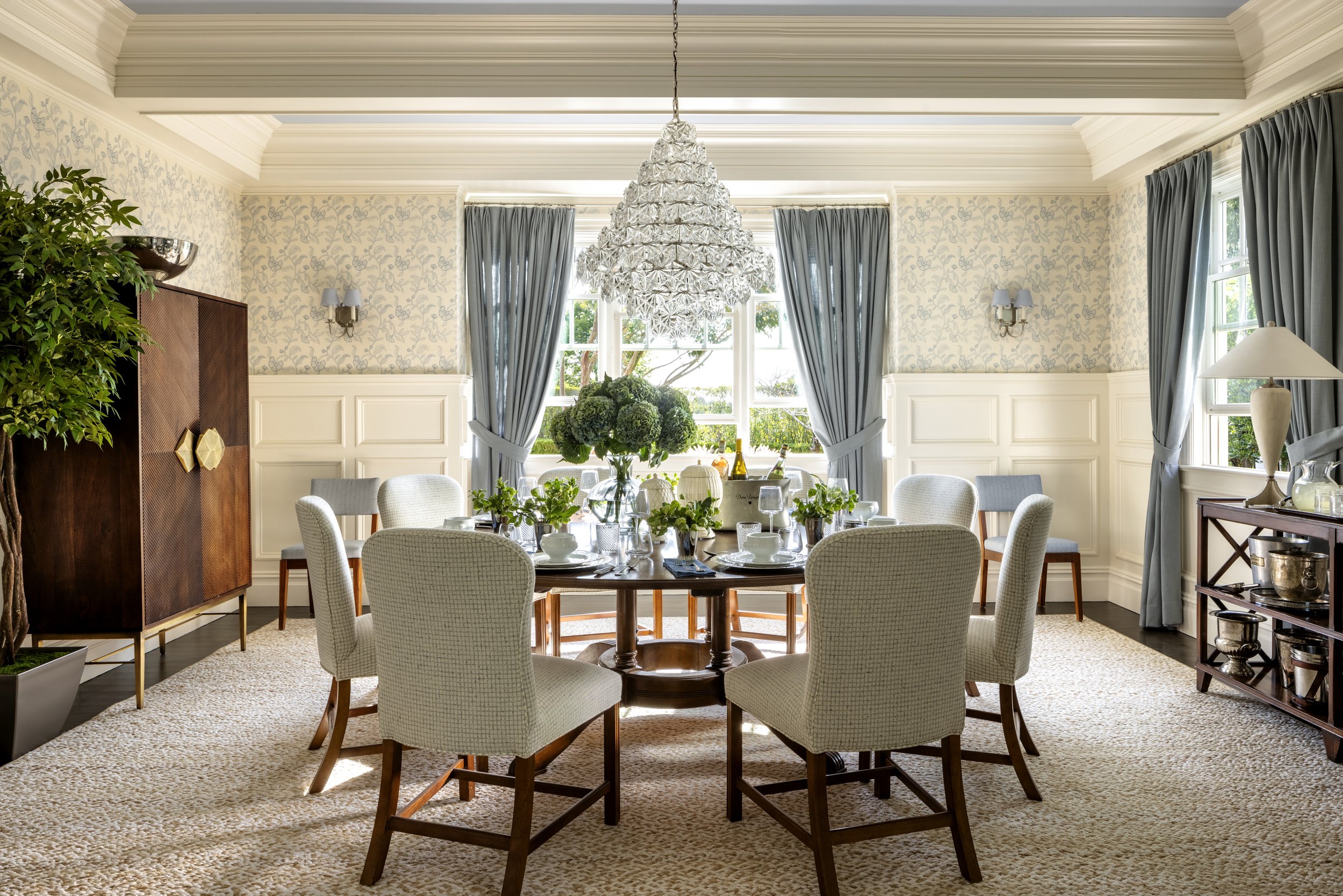 A dining room with a round wood table set with white dishes, glasses, and green plants. Beige chairs surround the table. Large window with blue curtains, a chandelier hangs above, and sunlight lets in through the window.